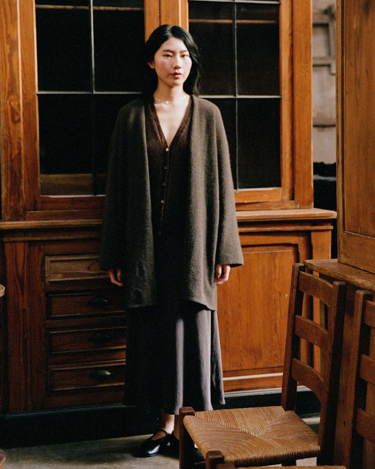 A woman stands indoors before a wooden cabinet, wearing the Cocoon Coat – Truffle in an oversized fit over a dark dress and black shoes. Wooden chairs appear in the foreground.