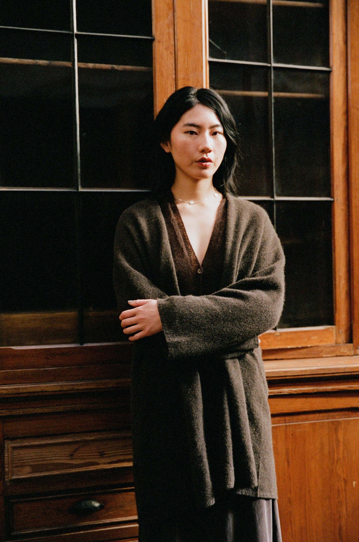 A woman with long black hair stands indoors in front of a wooden cabinet with dark glass panels, wearing the Cocoon Coat in Truffle and a neutral expression.