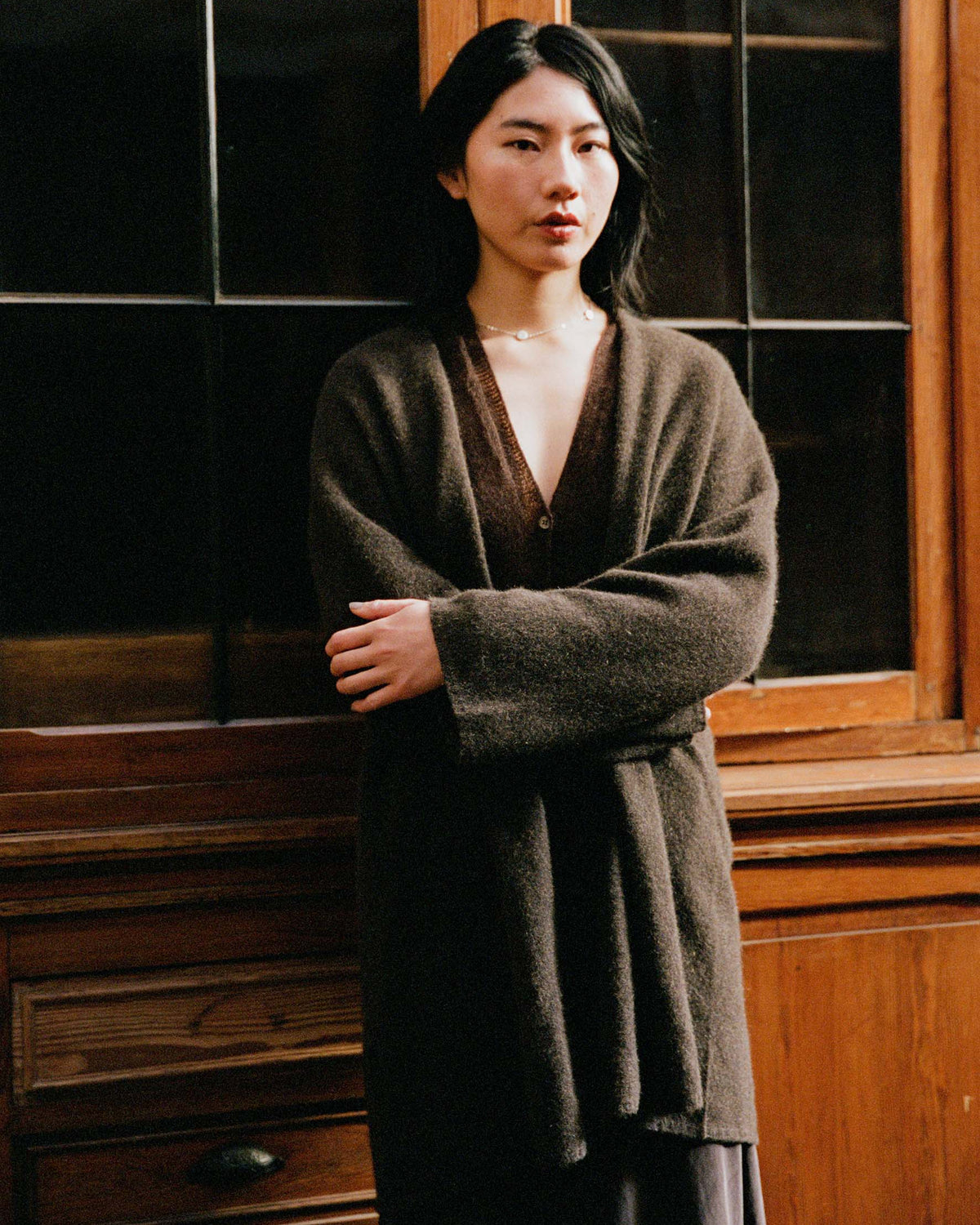 A woman with long black hair stands indoors in front of a wooden cabinet with dark glass panels, wearing the Cocoon Coat in Truffle and a neutral expression.
