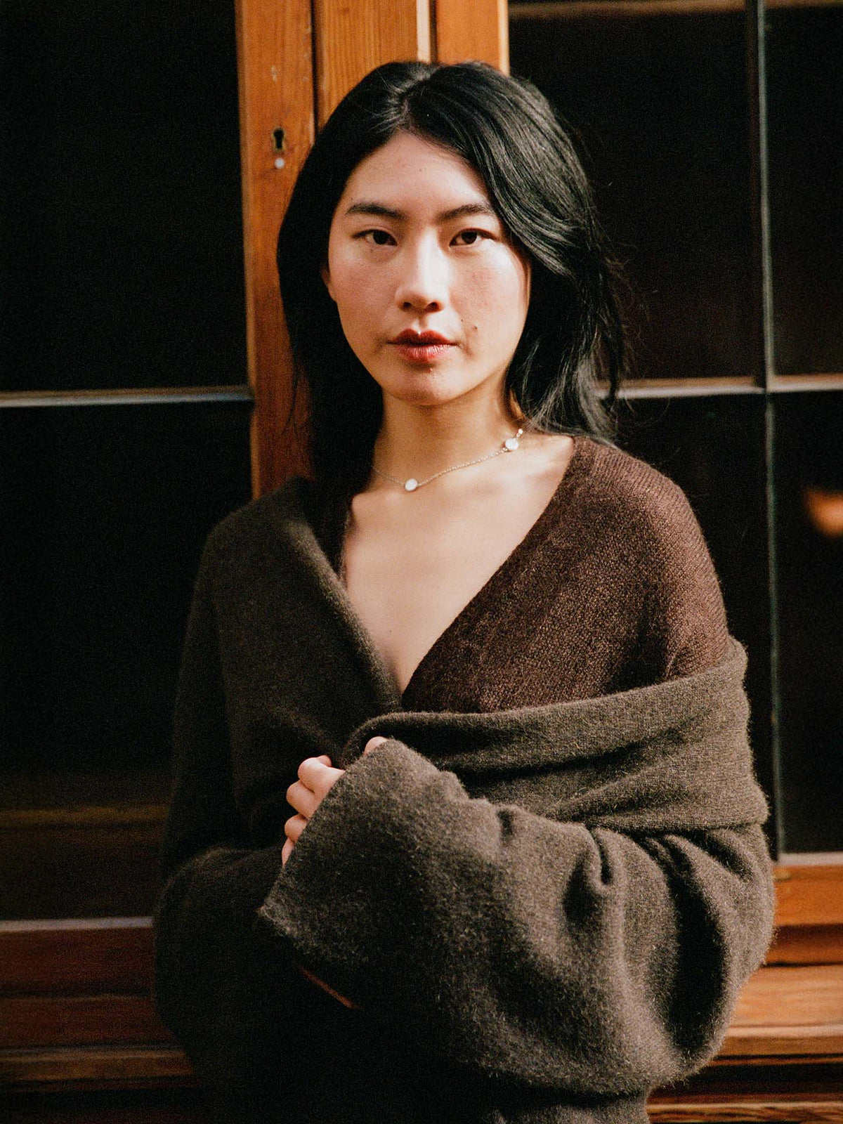 A woman with long black hair wears the Cocoon Coat in Truffle, standing indoors by a wooden-framed window and looking directly at the camera.