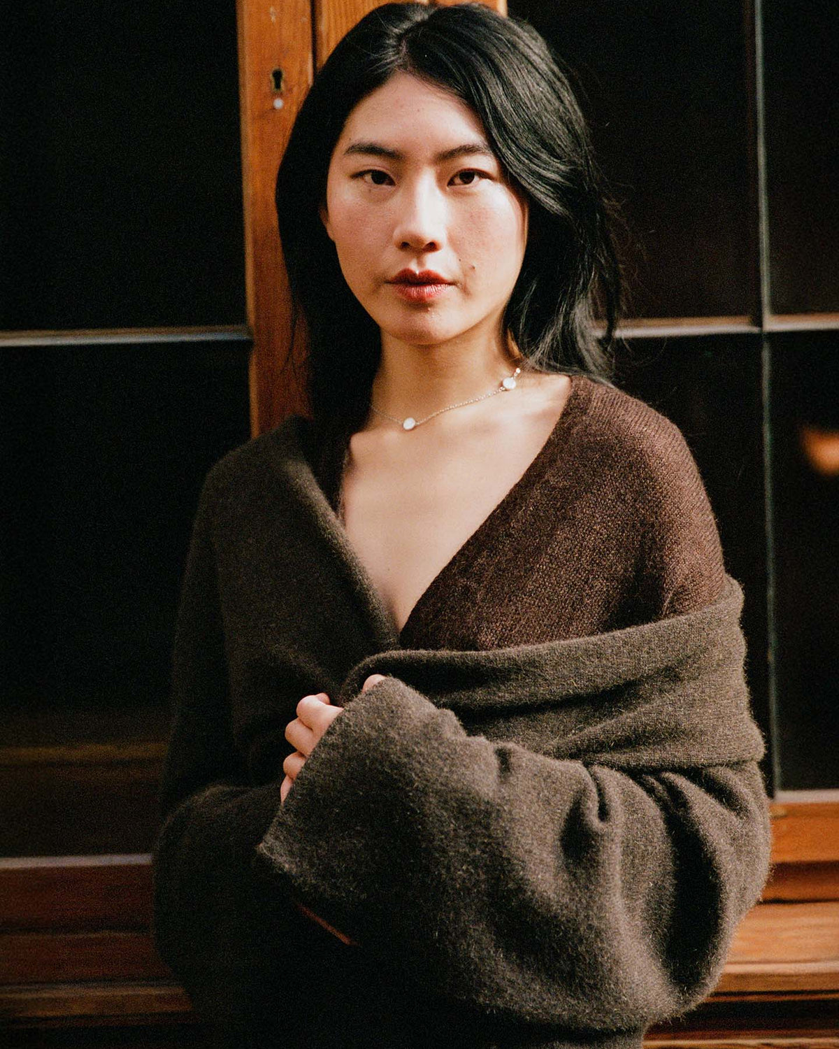 A woman with long black hair wears the Cocoon Coat in Truffle, standing indoors by a wooden-framed window and looking directly at the camera.