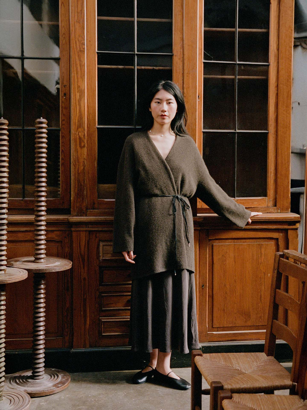 A woman stands indoors in front of wooden cabinets, wearing the Cocoon Coat – Truffle in an oversized style with a long skirt and black flats, one hand resting on the cabinet.