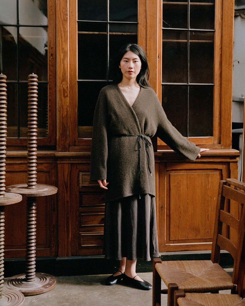A woman stands indoors in front of wooden cabinets, wearing the Cocoon Coat – Truffle in an oversized style with a long skirt and black flats, one hand resting on the cabinet.