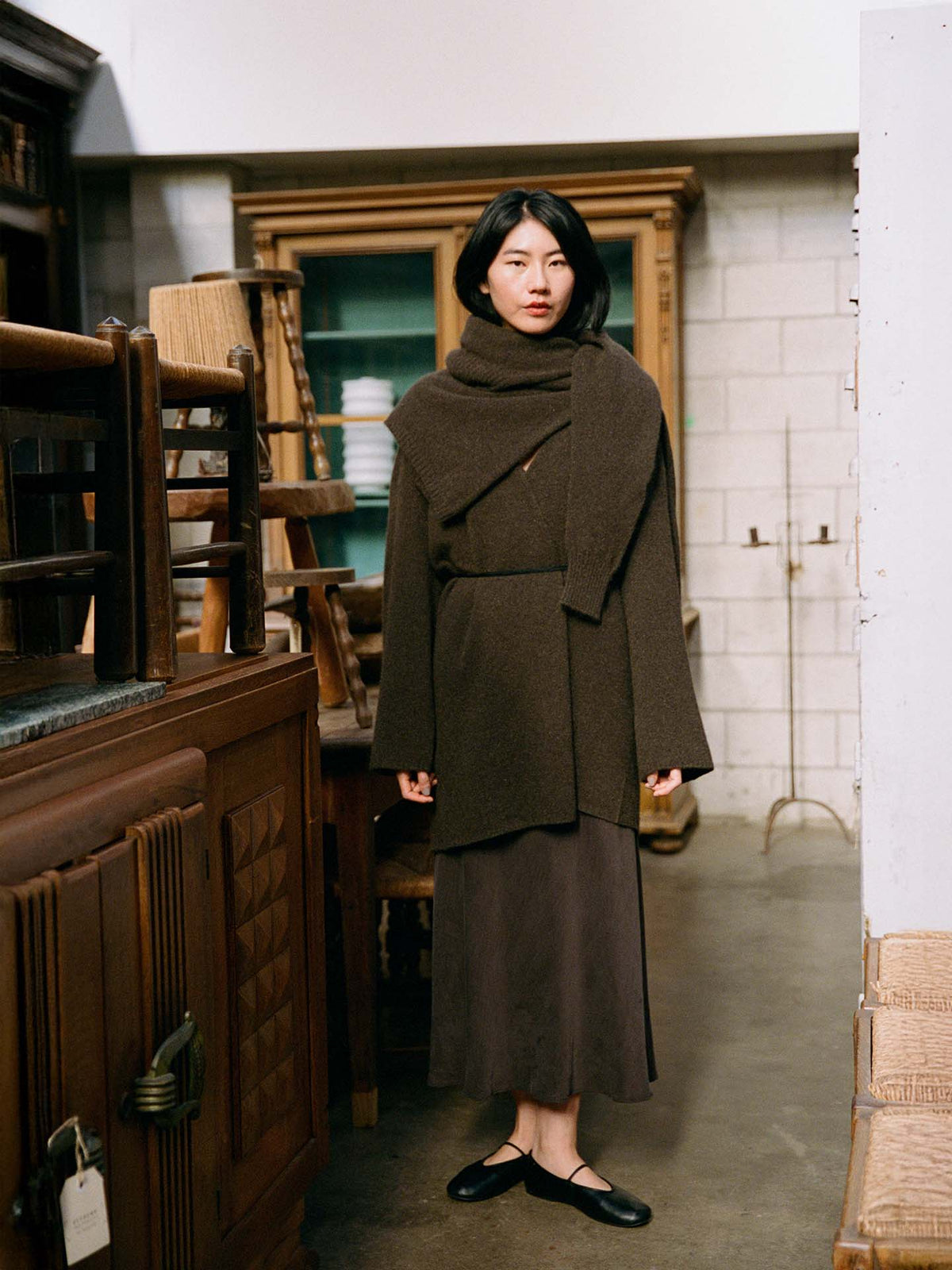A woman stands indoors wearing the Cocoon Coat – Truffle, an oversized dark brown wrap coat, paired with a long skirt and black shoes, surrounded by vintage wooden furniture and shelves.