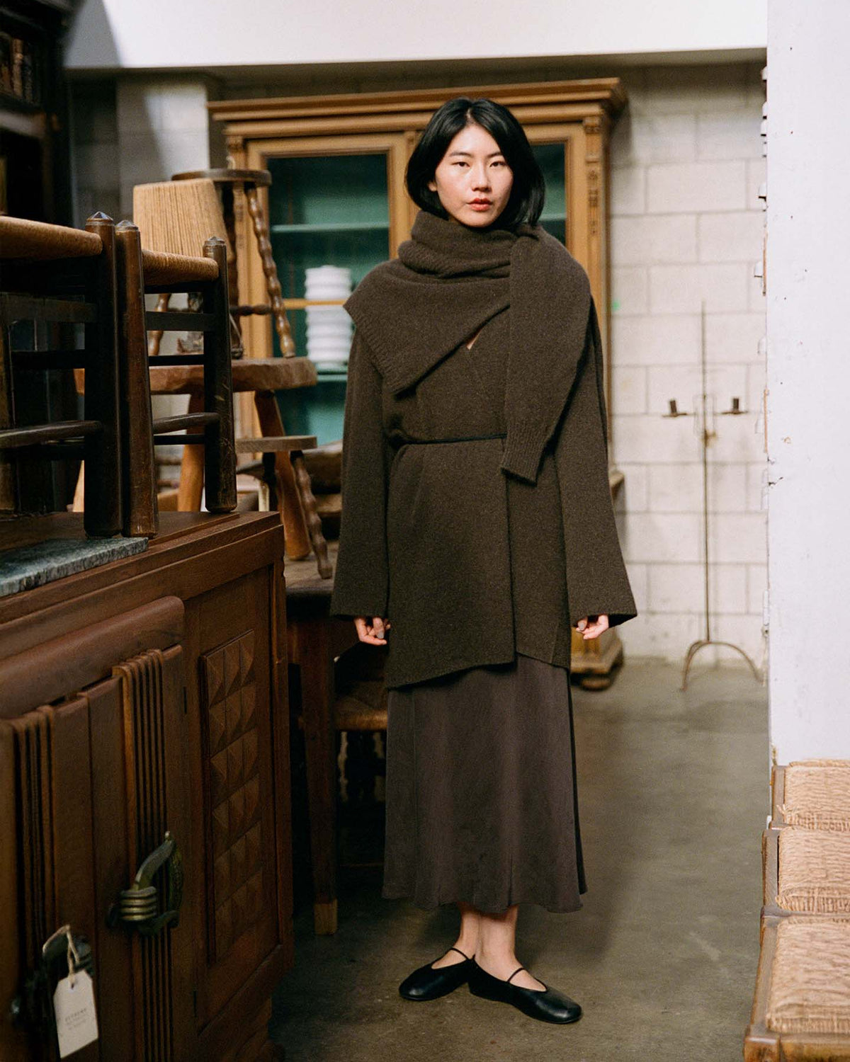A woman stands indoors wearing the Cocoon Coat – Truffle, an oversized dark brown wrap coat, paired with a long skirt and black shoes, surrounded by vintage wooden furniture and shelves.