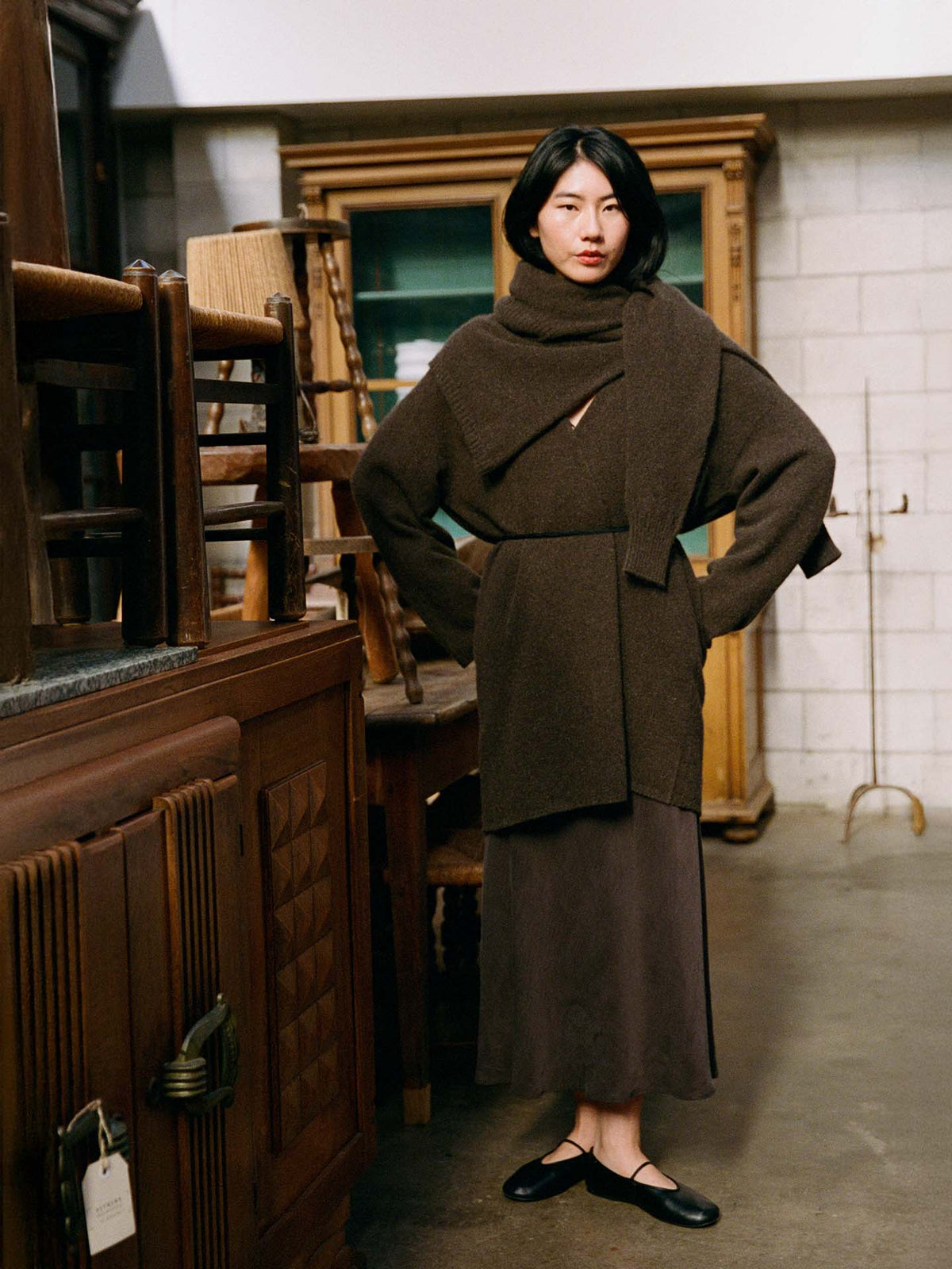 A woman stands indoors among wooden furniture, looking at the camera while wearing the Cocoon Coat – Truffle, a long brown skirt, and black flats.