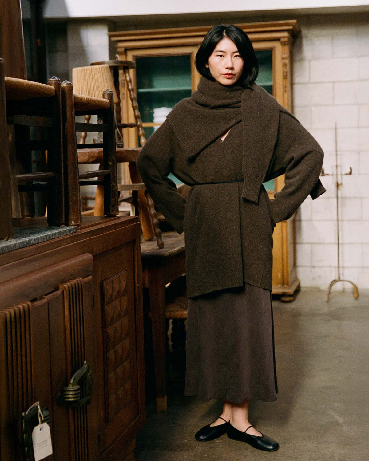 A woman stands indoors among wooden furniture, looking at the camera while wearing the Cocoon Coat – Truffle, a long brown skirt, and black flats.