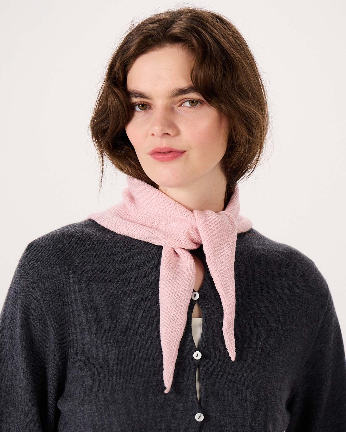 A woman with shoulder-length brown hair stands against a plain light background, wearing a dark gray button-up top and the Daisy Scarf in Barely Pink tied around her neck, with an extra wide yoga mat neatly rolled at her feet.
