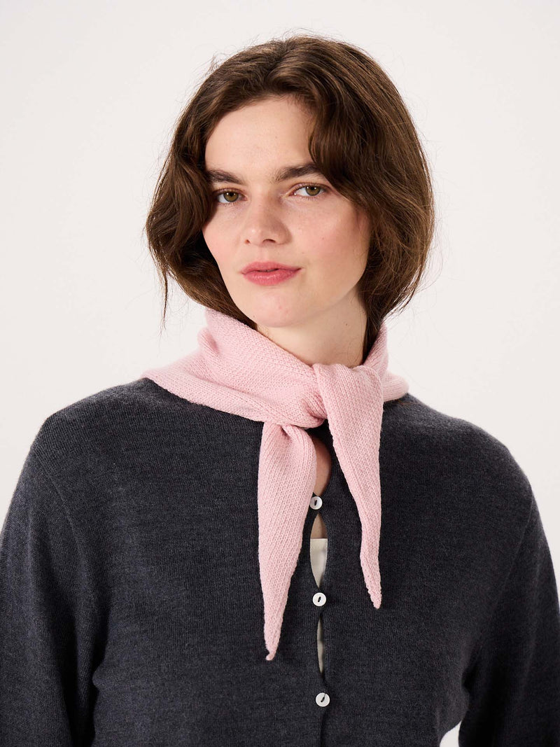 A woman with shoulder-length brown hair stands against a plain light background, wearing a dark gray button-up top and the Daisy Scarf in Barely Pink tied around her neck, with an extra wide yoga mat neatly rolled at her feet.