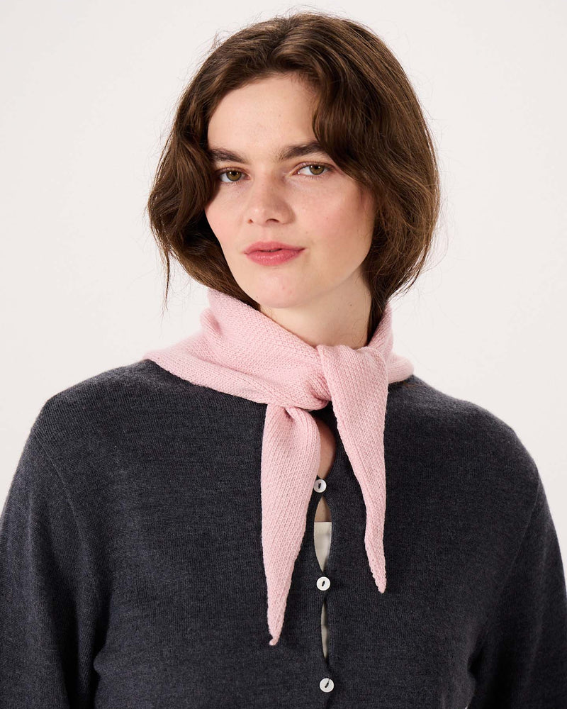 A woman with shoulder-length brown hair stands against a plain light background, wearing a dark gray button-up top and the Daisy Scarf in Barely Pink tied around her neck, with an extra wide yoga mat neatly rolled at her feet.
