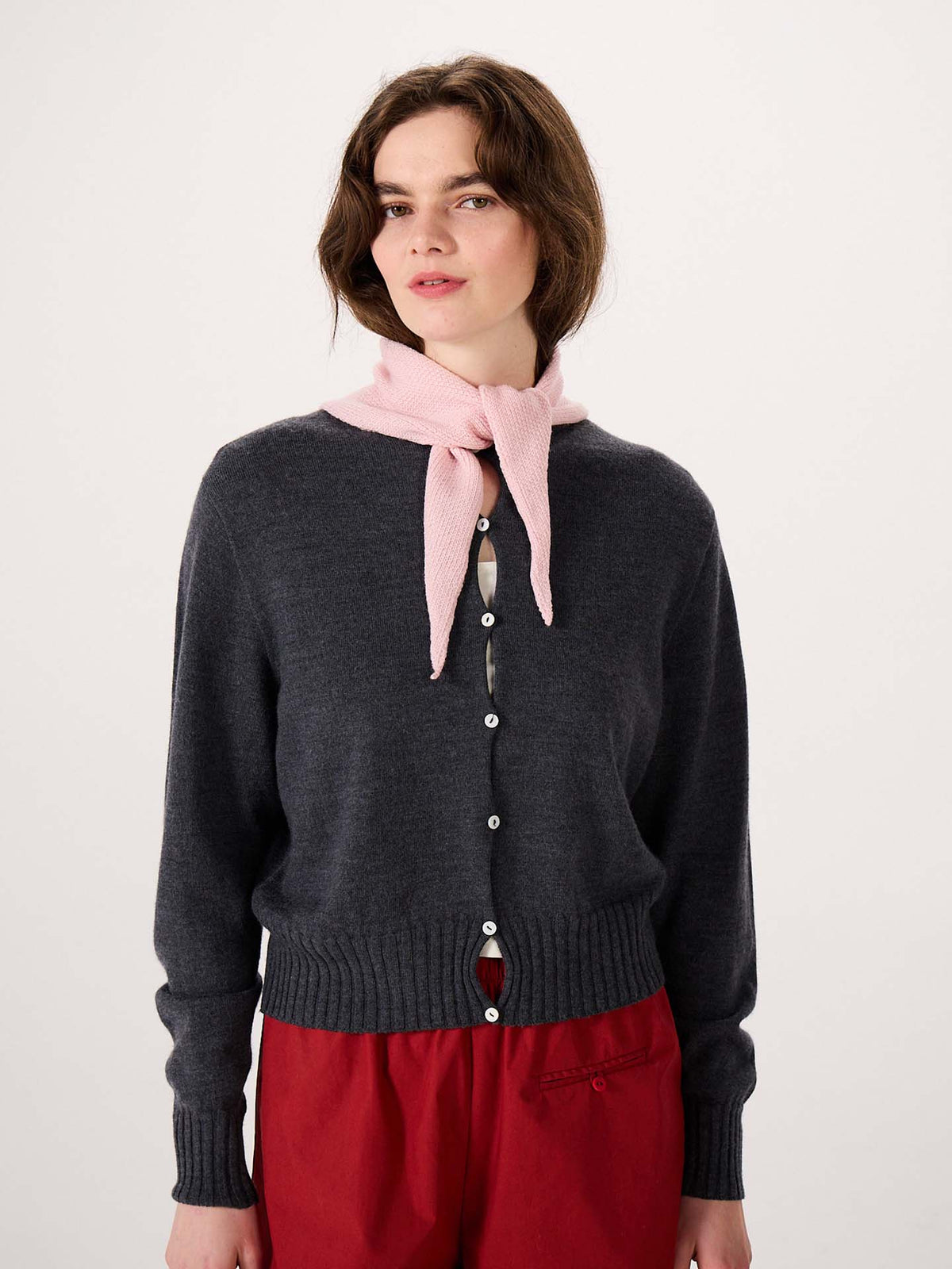 A person wearing a charcoal gray buttoned cardigan with the Daisy Scarf in Barely Pink around their neck and red pants stands on an extra wide yoga mat against a plain light background.