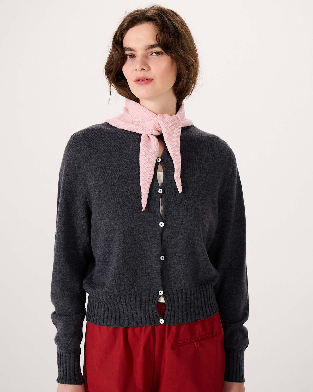 A person wearing a charcoal gray buttoned cardigan with the Daisy Scarf in Barely Pink around their neck and red pants stands on an extra wide yoga mat against a plain light background.