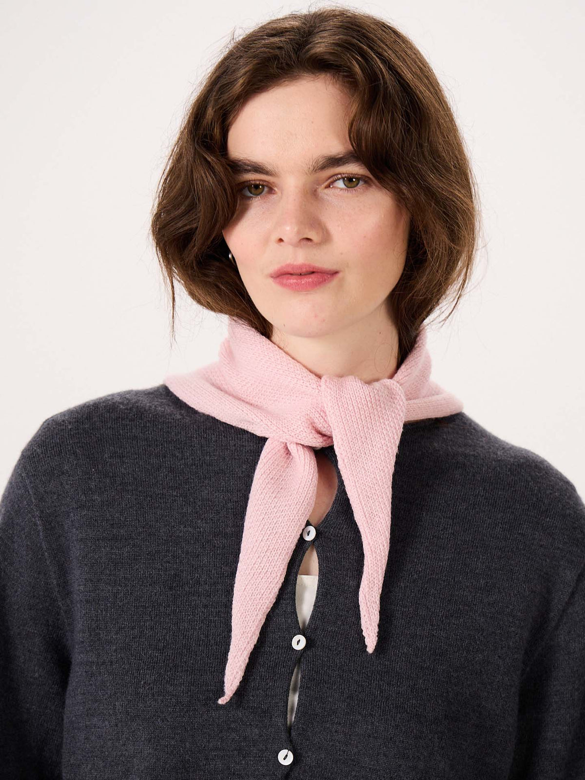 A woman wearing a dark sweater with large buttons and the Daisy Scarf in Barely Pink tied around her neck looks at the camera against a plain background, relaxing after her session on an extra wide yoga mat.
