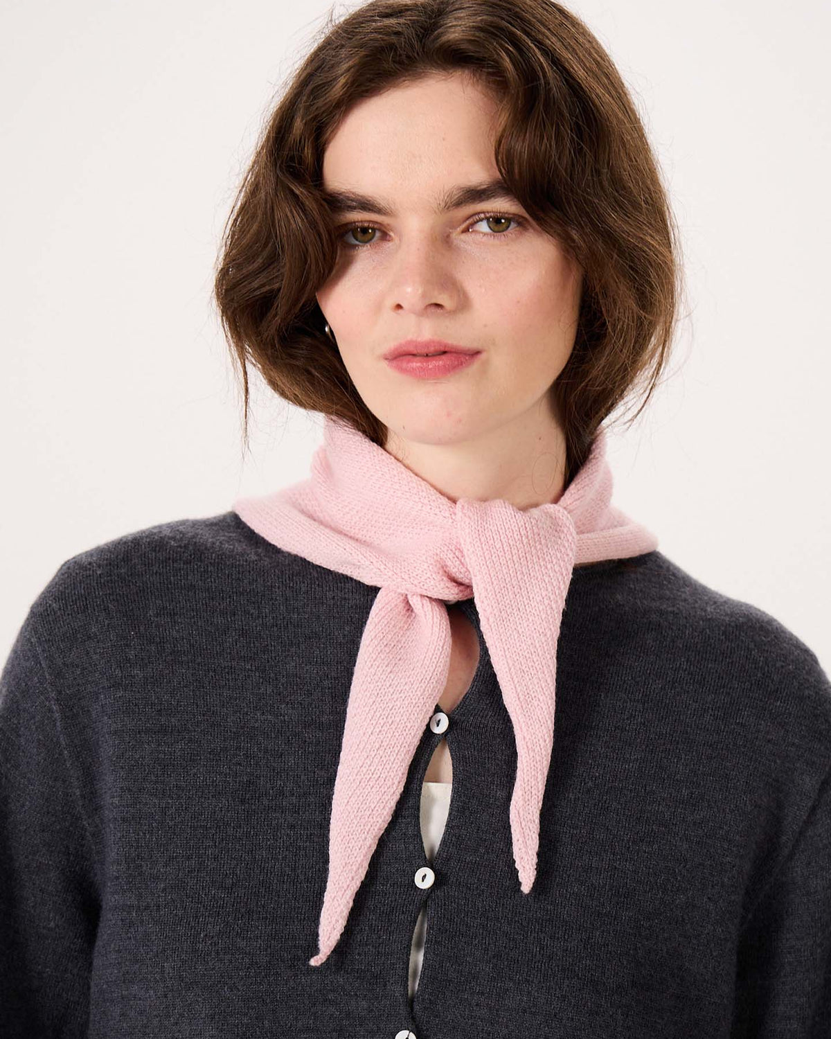 A woman wearing a dark sweater with large buttons and the Daisy Scarf in Barely Pink tied around her neck looks at the camera against a plain background, relaxing after her session on an extra wide yoga mat.