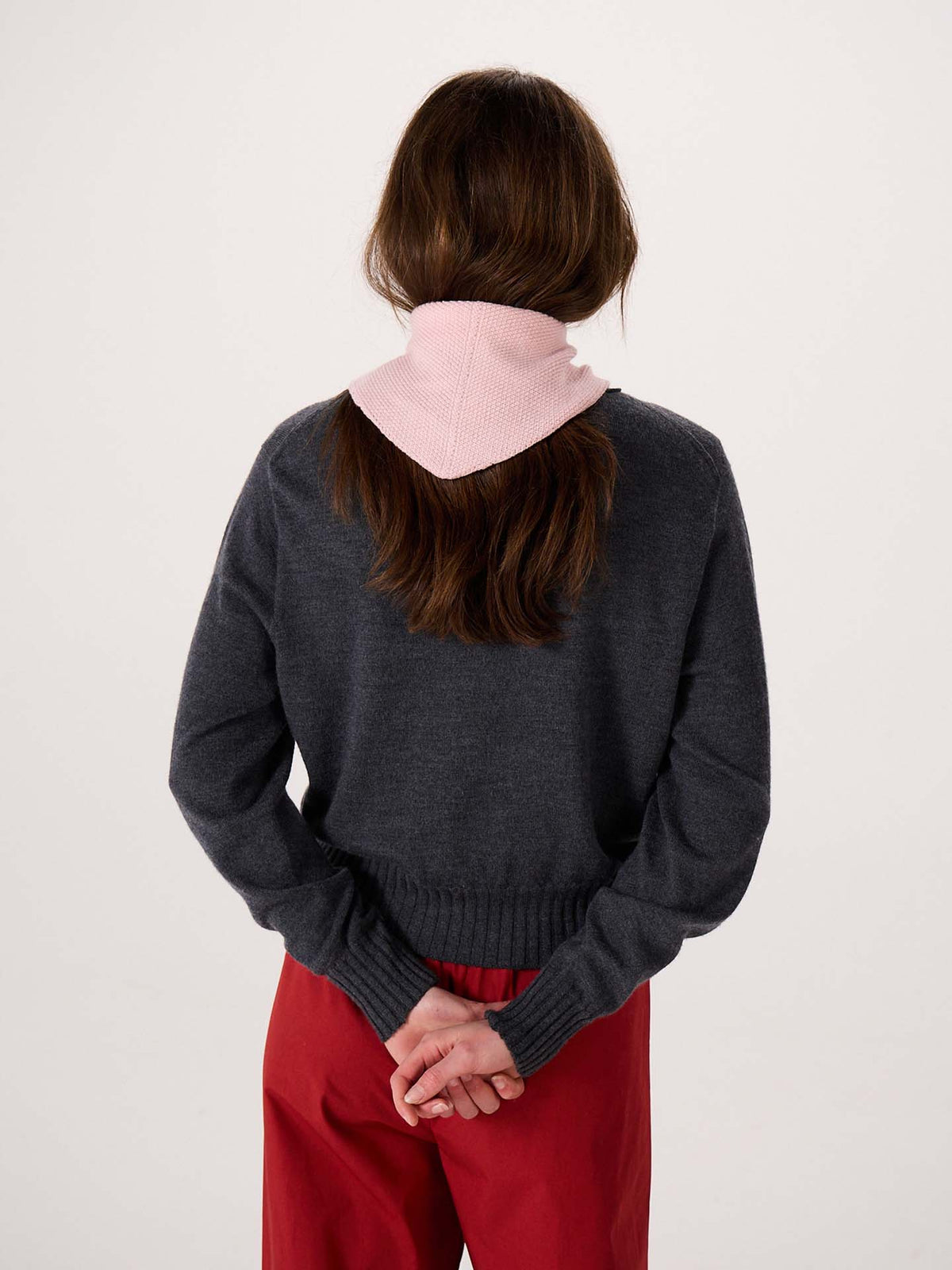 A person with long brown hair, wearing a gray sweater, red pants, and the Daisy Scarf – Barely Pink, stands facing away with hands clasped behind their back against a plain background, beside an extra wide yoga mat.