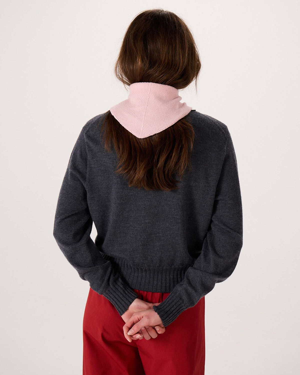 A person with long brown hair, wearing a gray sweater, red pants, and the Daisy Scarf – Barely Pink, stands facing away with hands clasped behind their back against a plain background, beside an extra wide yoga mat.
