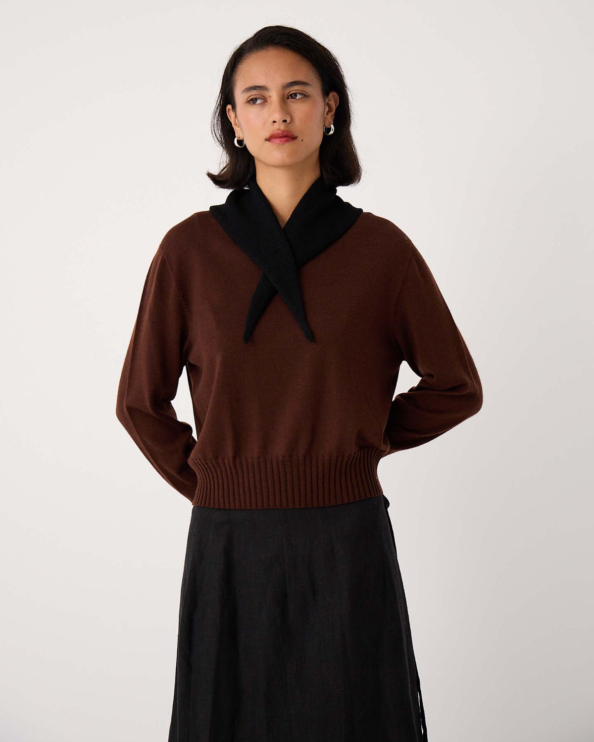 A woman stands against a plain background, wearing a brown sweater and dark skirt with the Daisy Scarf – Black draped over her shoulders (97cm long, 22cm wide), styled to display its size as she looks slightly to the side.