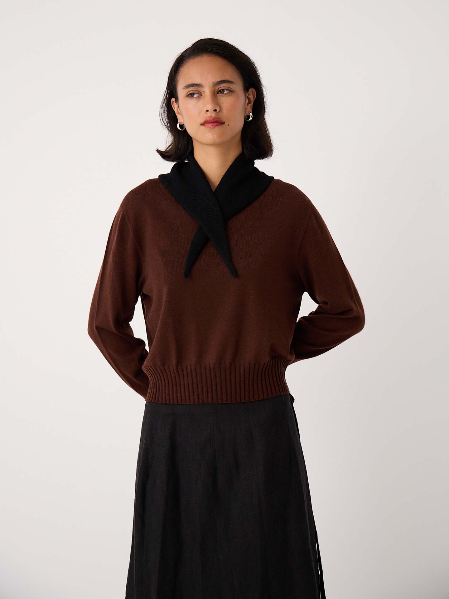 A woman stands against a plain background, wearing a brown sweater and dark skirt with the Daisy Scarf – Black draped over her shoulders (97cm long, 22cm wide), styled to display its size as she looks slightly to the side.