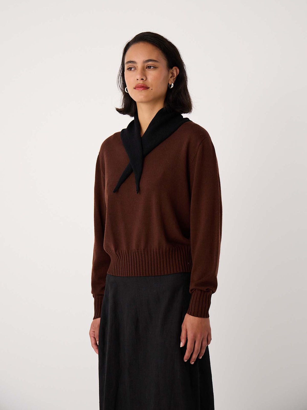 A woman stands against a plain background wearing a brown sweater, a long black skirt, and the Daisy Scarf – Black (22cm wide, 97cm long) draped around her neck.