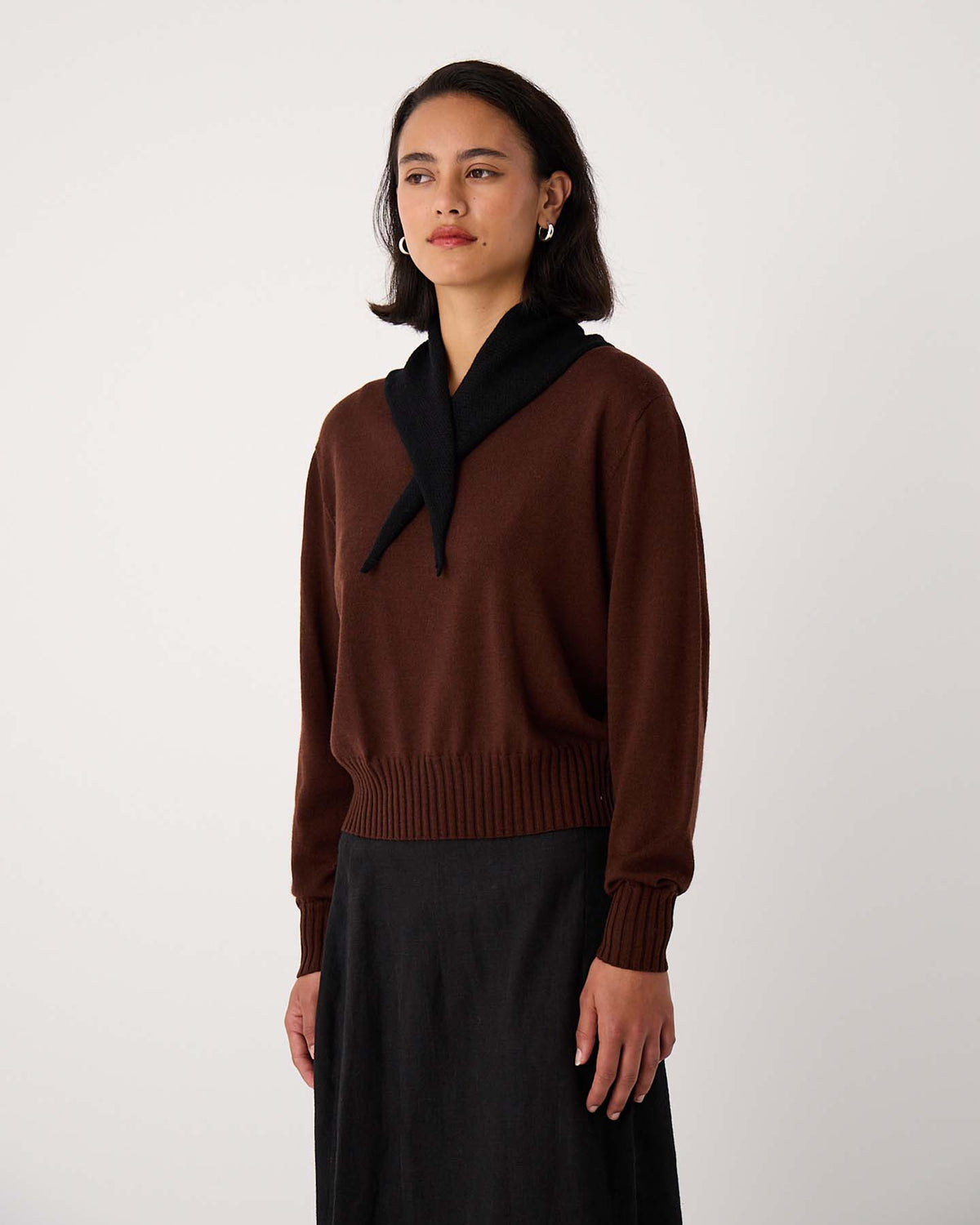 A woman stands against a plain background wearing a brown sweater, a long black skirt, and the Daisy Scarf – Black (22cm wide, 97cm long) draped around her neck.