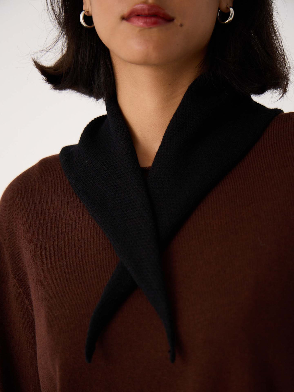 A woman wears a brown top with the Daisy Scarf – Black draped around her neck and silver hoop earrings, shown from shoulders to lips—perfectly styled for her next workout on a 97cm yoga mat.