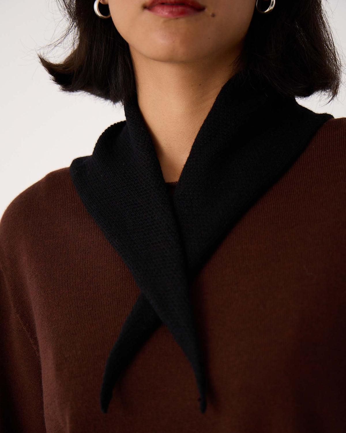 A woman wears a brown top with the Daisy Scarf – Black draped around her neck and silver hoop earrings, shown from shoulders to lips—perfectly styled for her next workout on a 97cm yoga mat.