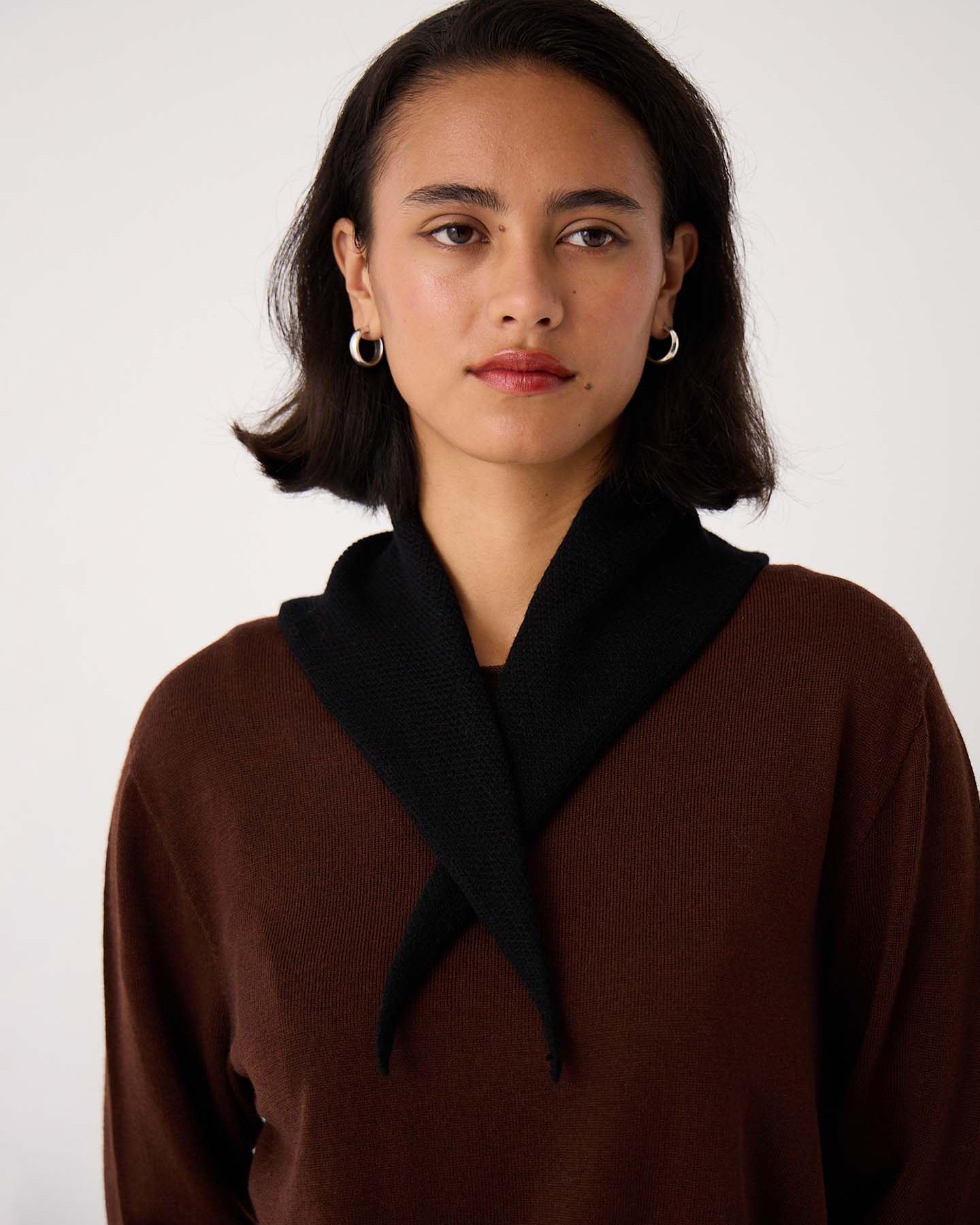 A woman with short dark hair and hoop earrings wears a brown top and the Daisy Scarf – Black (97cm x 22cm), standing against a plain light background.