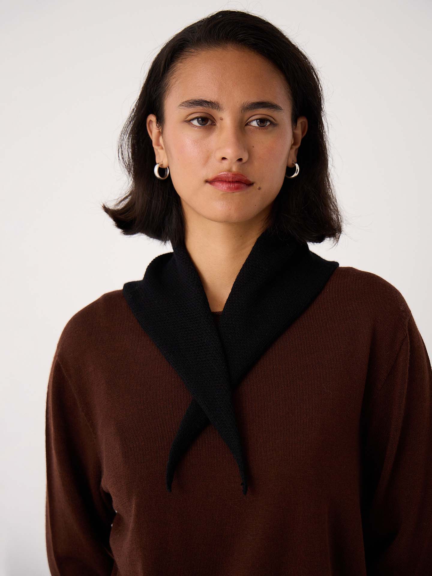 A woman with short dark hair and hoop earrings wears a brown top and the Daisy Scarf – Black (97cm x 22cm), standing against a plain light background.