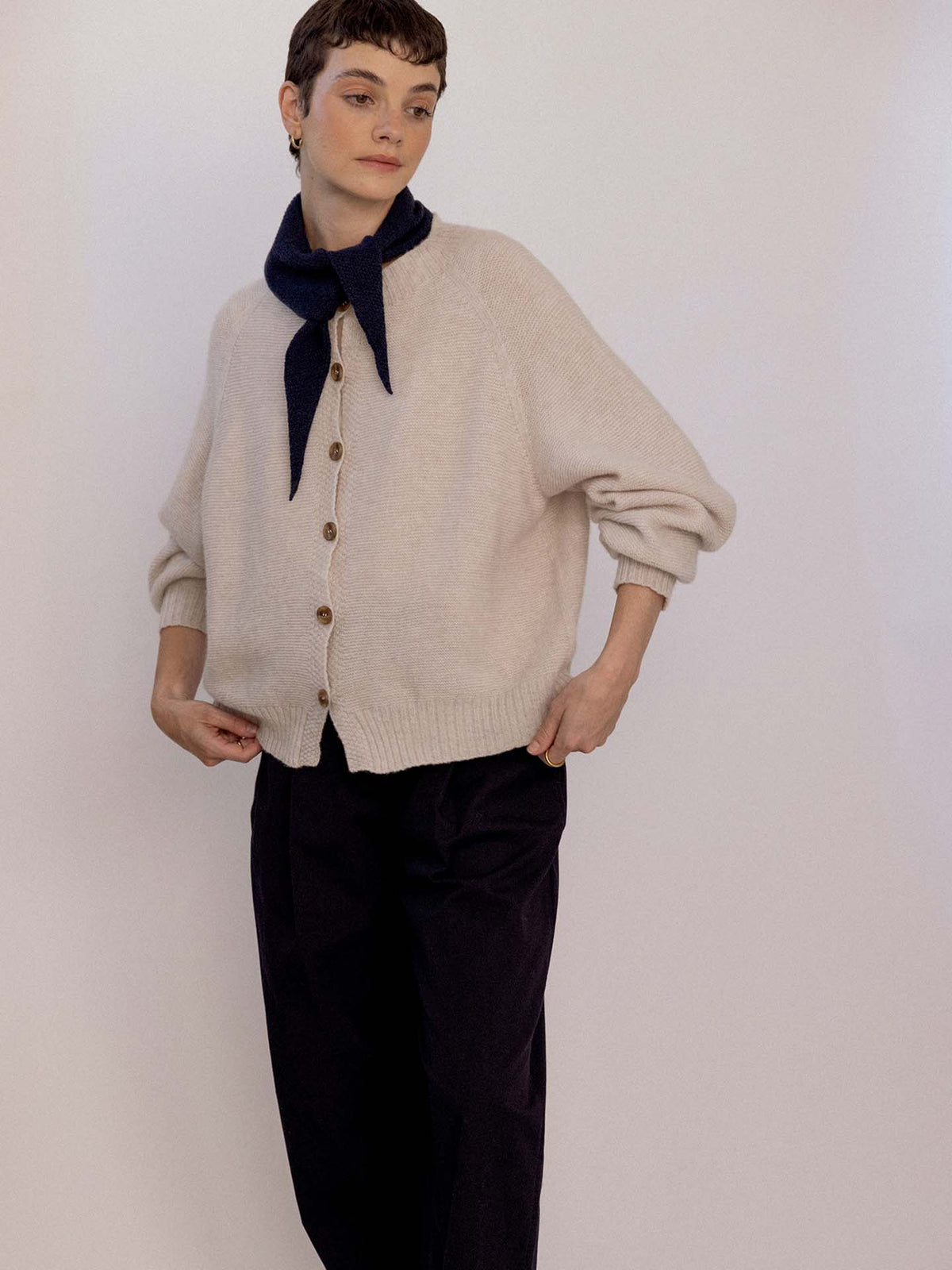 A person wearing a beige button-up cardigan, black pants, and the Daisy Scarf – Black tied around their neck stands against a plain light background beside an extra wide yoga mat.