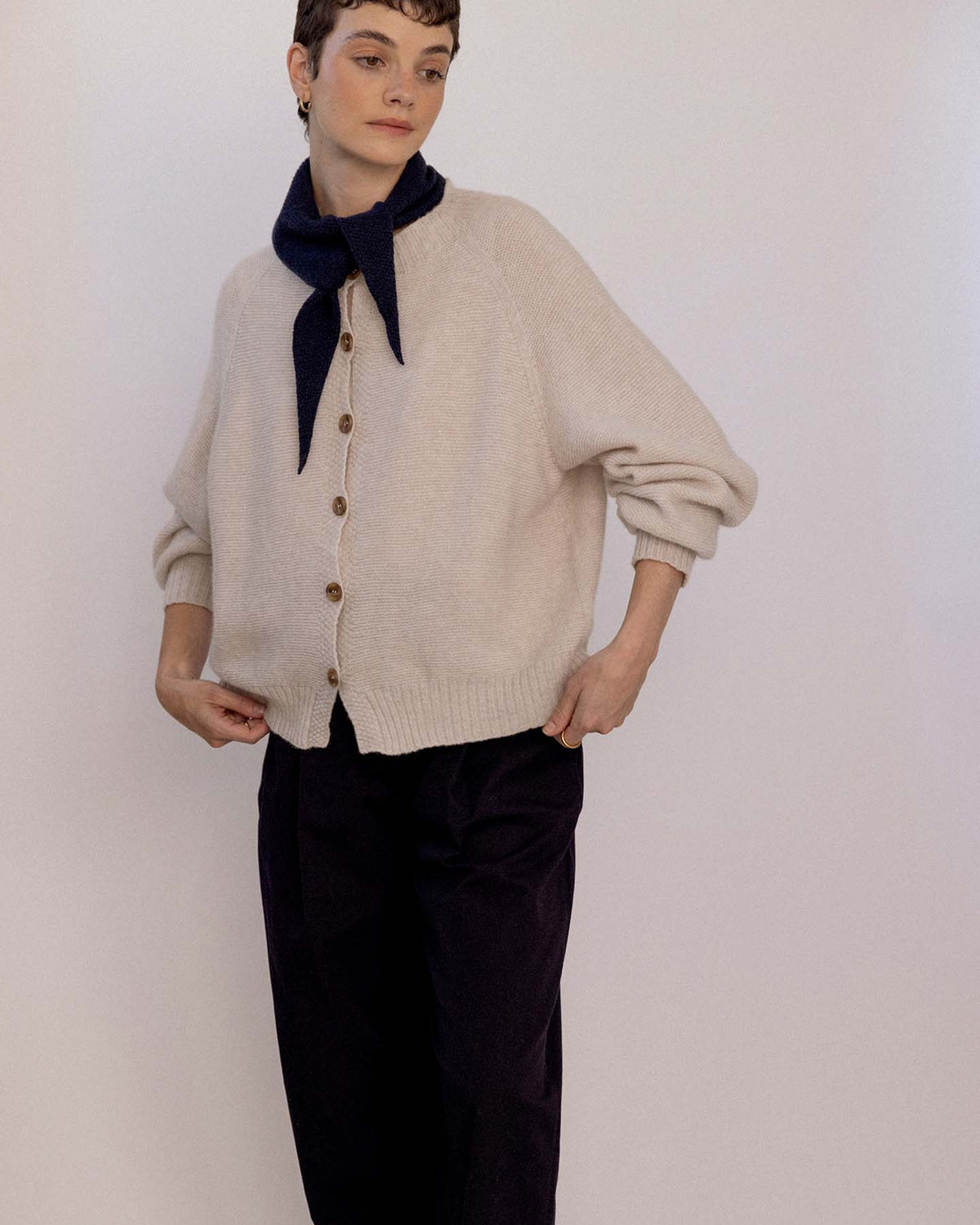 A person wearing a beige button-up cardigan, black pants, and the Daisy Scarf – Black tied around their neck stands against a plain light background beside an extra wide yoga mat.