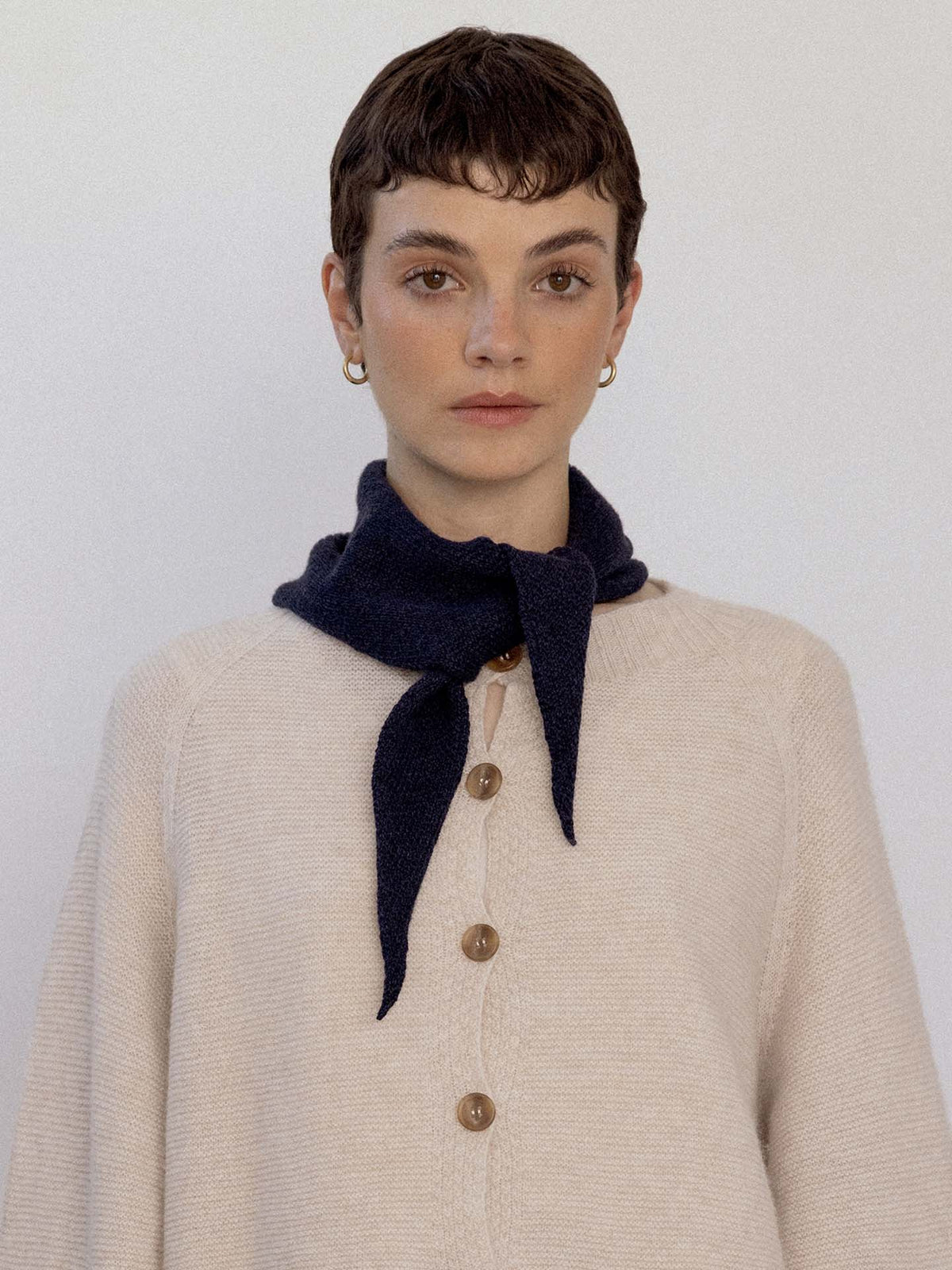 Person with short brown hair wears a cream buttoned sweater and the Daisy Scarf – Black, tied around the neck, standing against a plain light background next to an extra wide yoga mat.