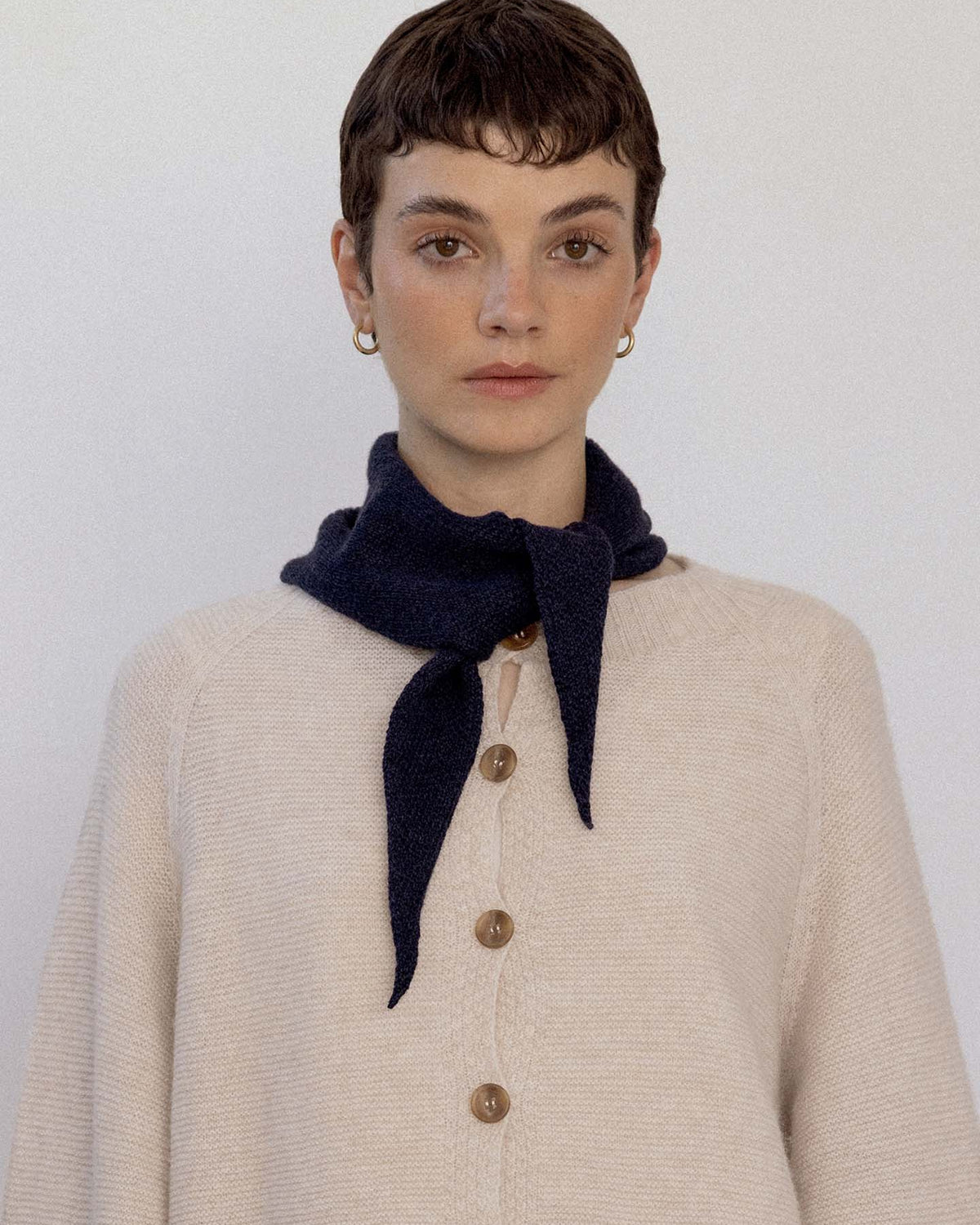 Person with short brown hair wears a cream buttoned sweater and the Daisy Scarf – Black, tied around the neck, standing against a plain light background next to an extra wide yoga mat.