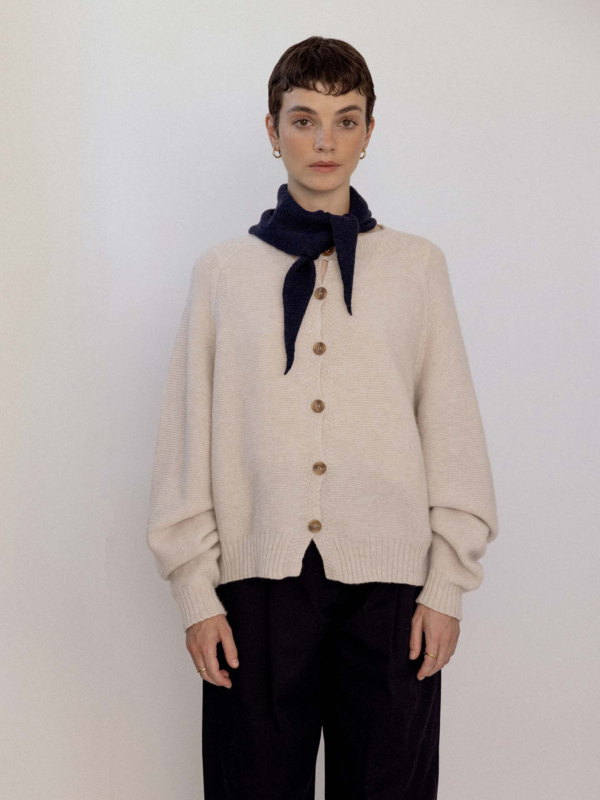 A person stands against a plain background, wearing a beige buttoned cardigan, black pants, and the Daisy Scarf – Black draped over their shoulders and tied at the neck, next to an extra wide yoga mat neatly rolled at their feet.