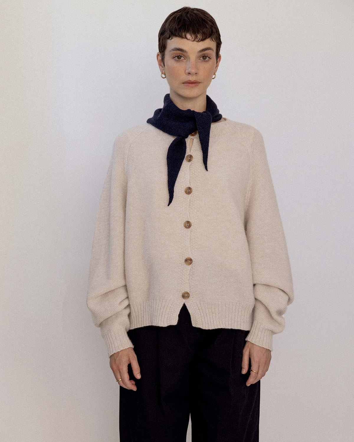 A person stands against a plain background, wearing a beige buttoned cardigan, black pants, and the Daisy Scarf – Black draped over their shoulders and tied at the neck, next to an extra wide yoga mat neatly rolled at their feet.