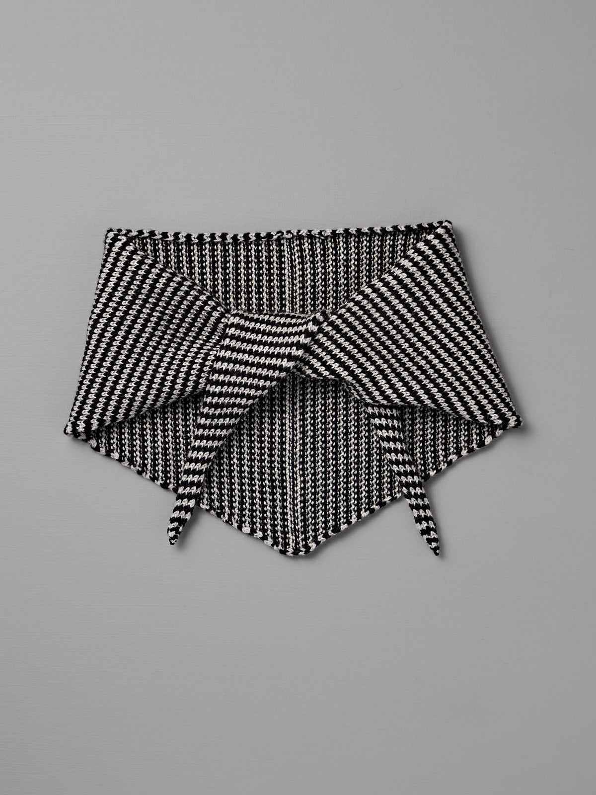The Daisy Scarf – Black Stripe by Francie is a black and white knitted neck warmer with a triangular shape and tie-front, shown flat on a gray background. This cozy accessory measures 22cm wide and 97cm long.