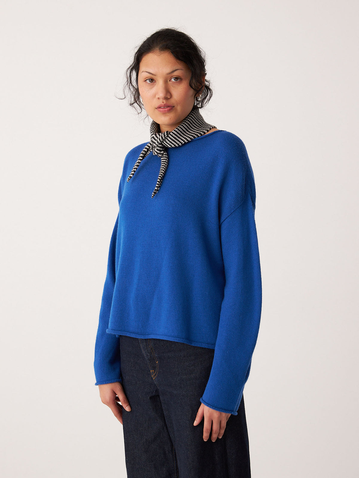 A woman faces forward against a plain light background, wearing a bright blue sweater, dark jeans, and the Francie Daisy Scarf – Black Stripe, a 97cm-long black-and-white striped neck scarf.