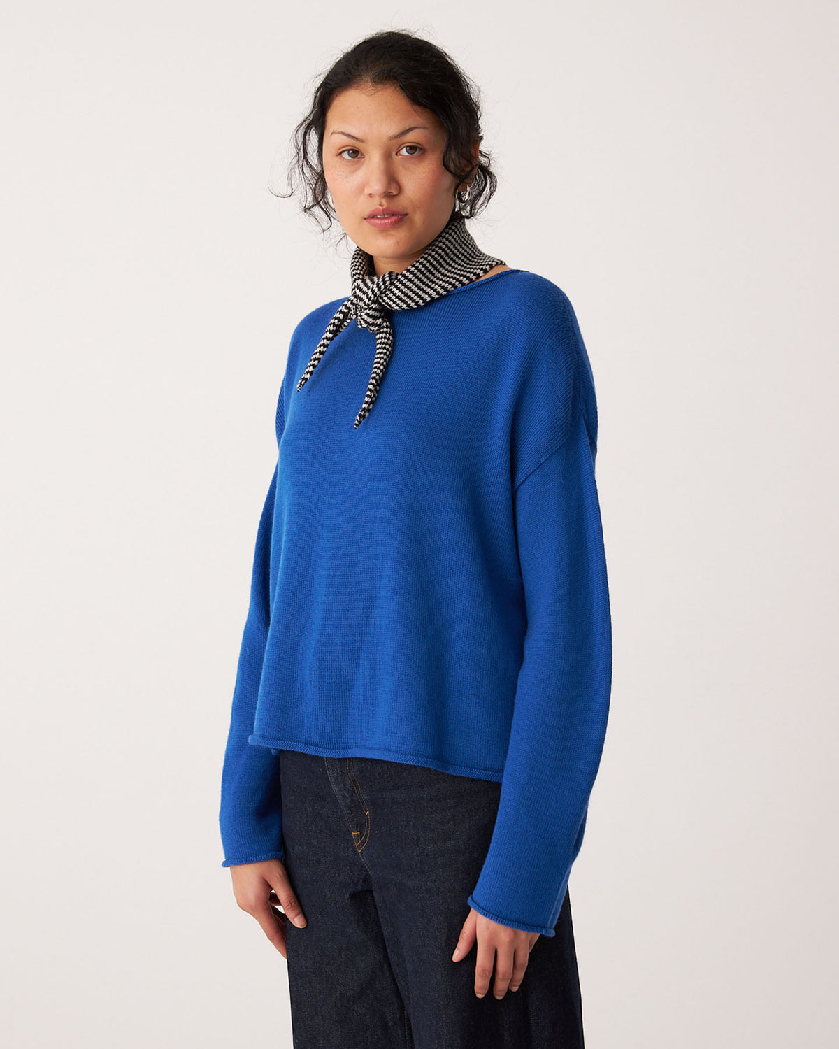 A person stands against a plain background wearing a bright blue sweater, dark jeans, and the Daisy Scarf – Black & Creme Stripe (97cm x 22cm).