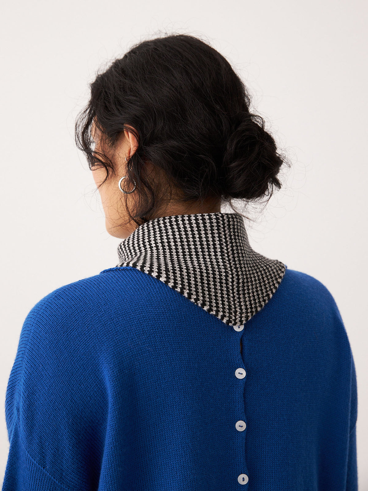 A person with dark hair in a low bun is facing away, wearing the Francie Daisy Scarf – Black Stripe (97cm x 22cm) over a blue sweater with back buttons and a black-and-white houndstooth turtleneck.