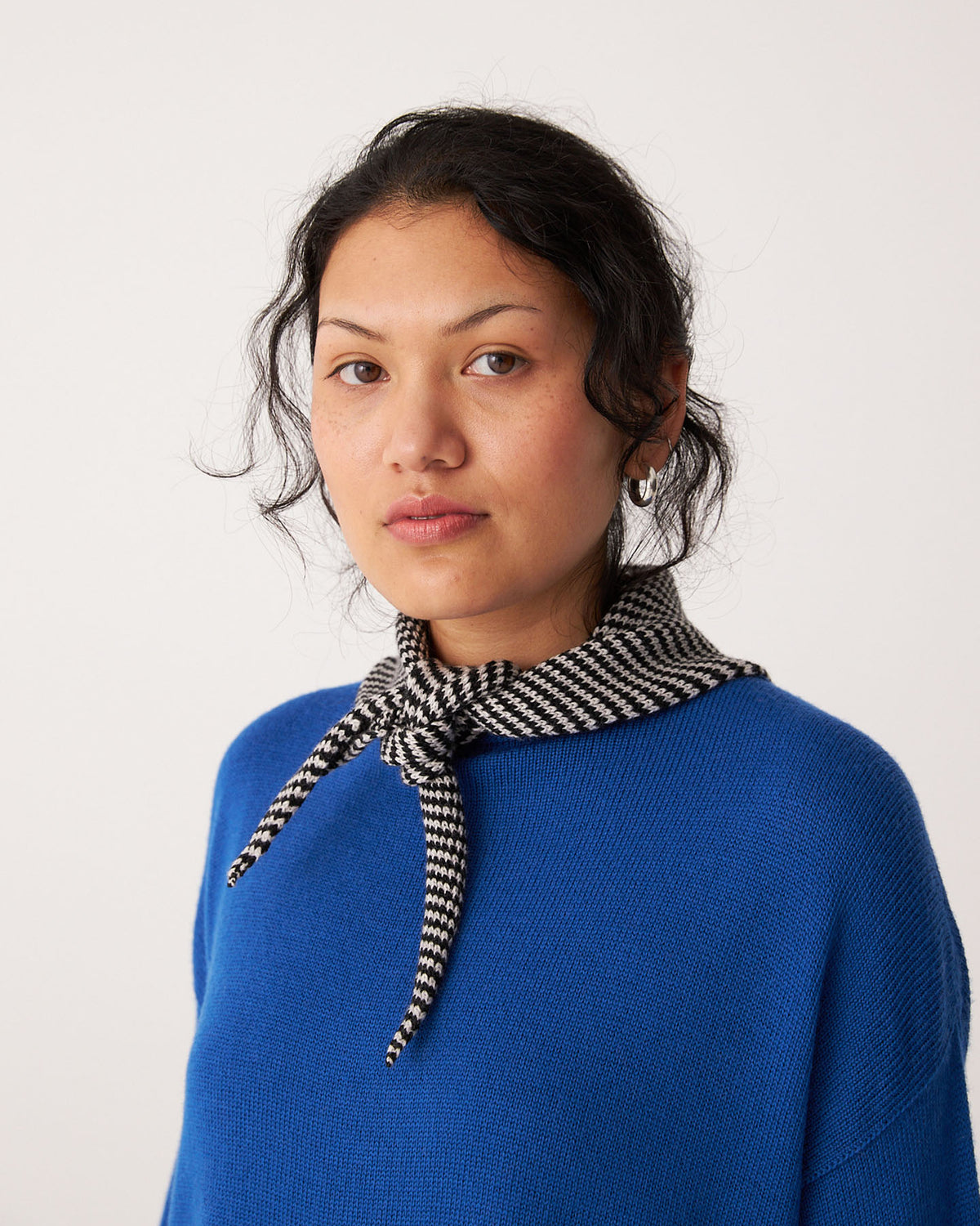 A person with dark hair wears a blue sweater and the Daisy Scarf – Black & Creme Stripe, measuring 22cm wide by 97cm long, looking at the camera against a plain background.