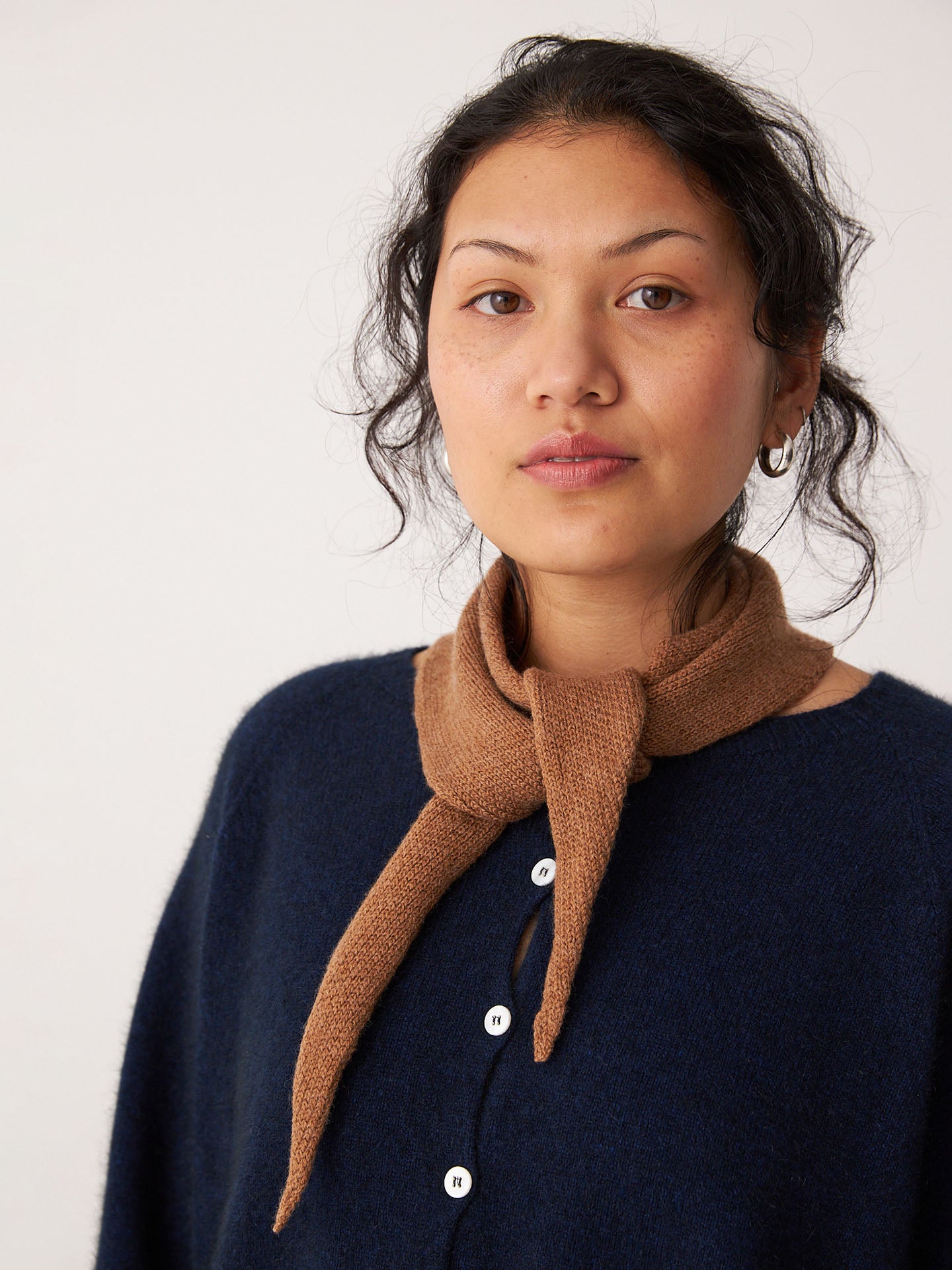 A person with wavy hair wears a dark blue buttoned top and the Francie Daisy Scarf – Brown Sugar (97cm x 22cm) tied around their neck, standing before a plain light background.