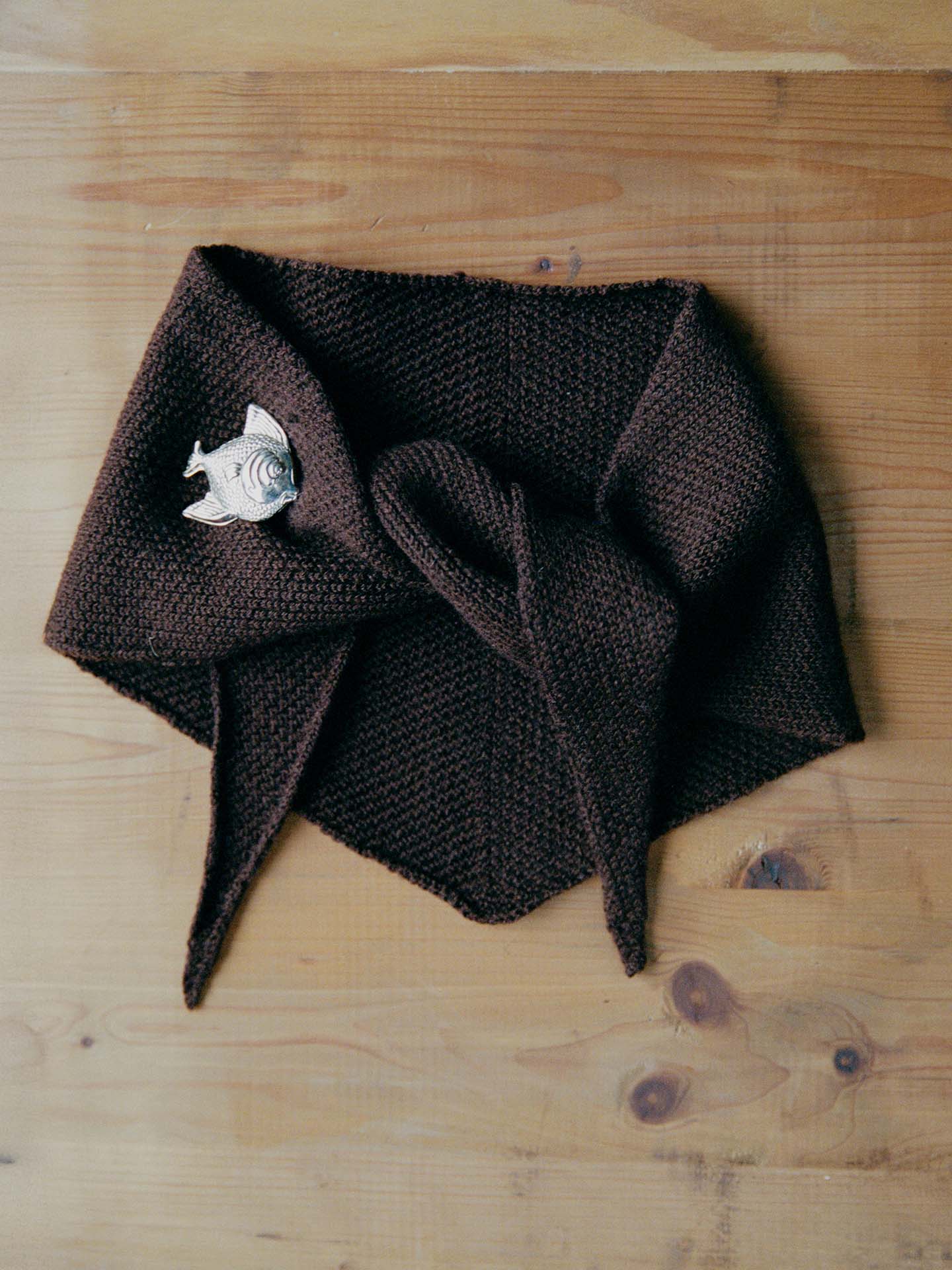 The Daisy Scarf – Cocoa, a brown knitted scarf with pointed ends, is displayed flat on a wooden surface and fastened with a silver fish-shaped brooch.