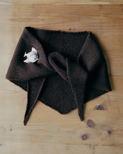 The Daisy Scarf – Cocoa, a brown knitted scarf with pointed ends, is displayed flat on a wooden surface and fastened with a silver fish-shaped brooch.