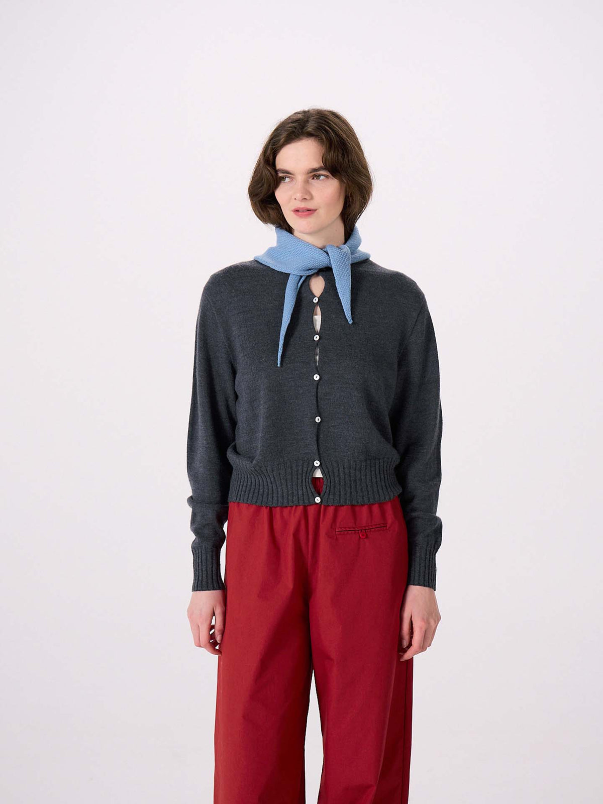 A woman stands against a plain white background, draping the Daisy Scarf – Celeste Blue over her shoulders. She wears a dark gray buttoned cardigan (97cm long and 22cm wide for a comfortable, tailored fit) and red pants.