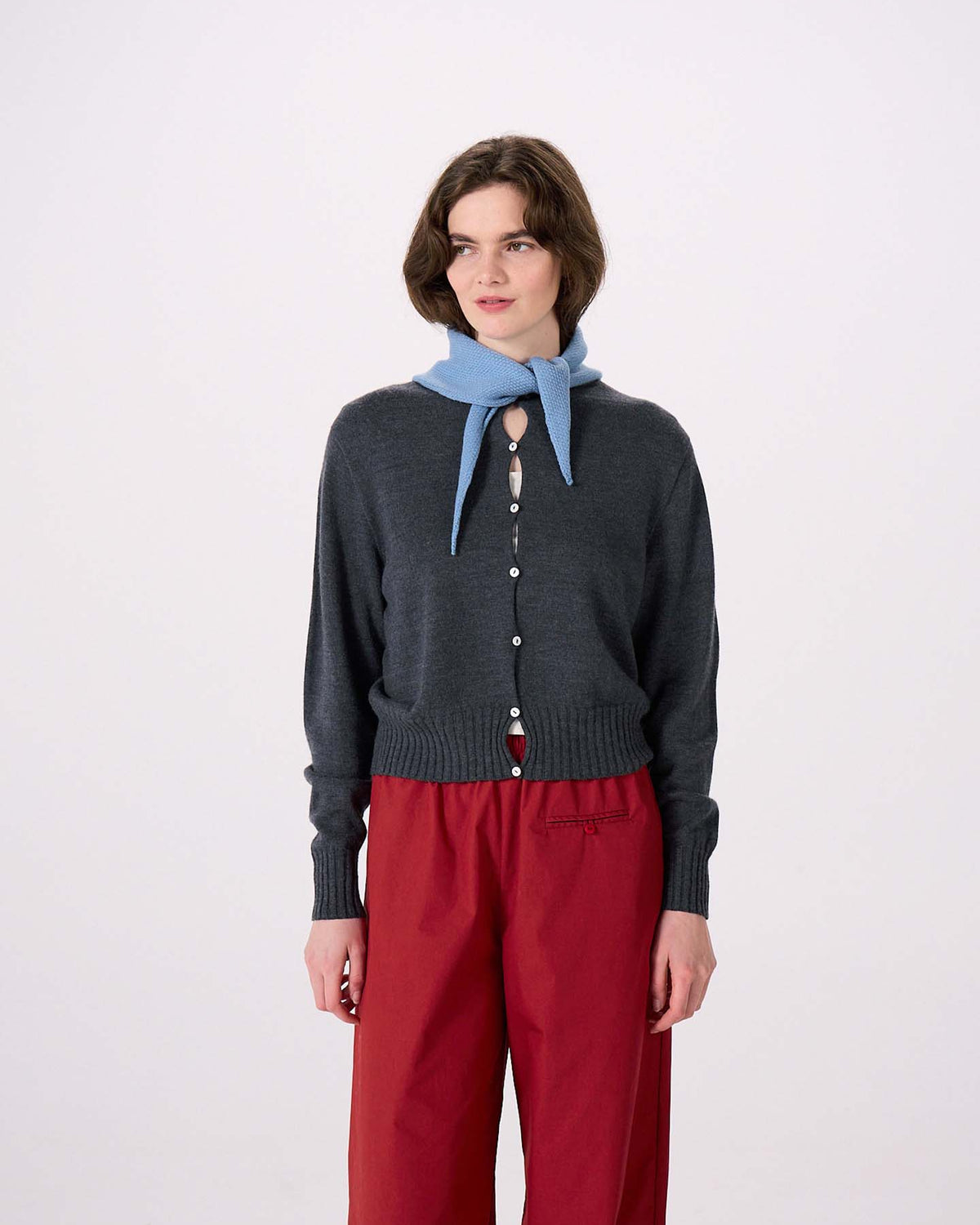 A woman stands against a plain white background, draping the Daisy Scarf – Celeste Blue over her shoulders. She wears a dark gray buttoned cardigan (97cm long and 22cm wide for a comfortable, tailored fit) and red pants.