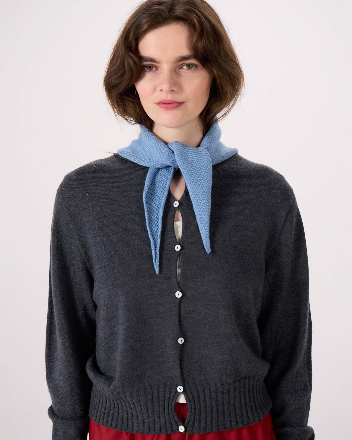 A woman with wavy brown hair wears a dark gray buttoned cardigan (97cm long by 22cm wide), red pants, and the Daisy Scarf – Celeste Blue tied around her neck, standing against a plain white background. The cardigan’s dimensions create an elegant silhouette.