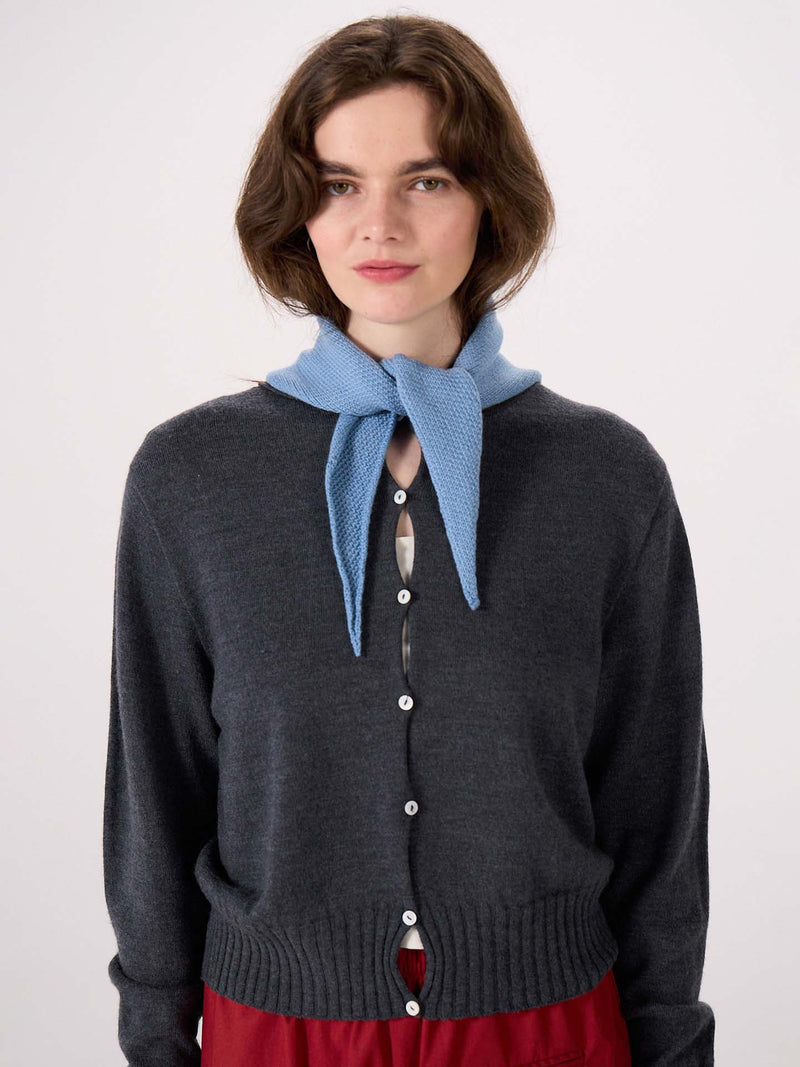 A woman with wavy brown hair wears a dark gray buttoned cardigan (97cm long by 22cm wide), red pants, and the Daisy Scarf – Celeste Blue tied around her neck, standing against a plain white background. The cardigan’s dimensions create an elegant silhouette.