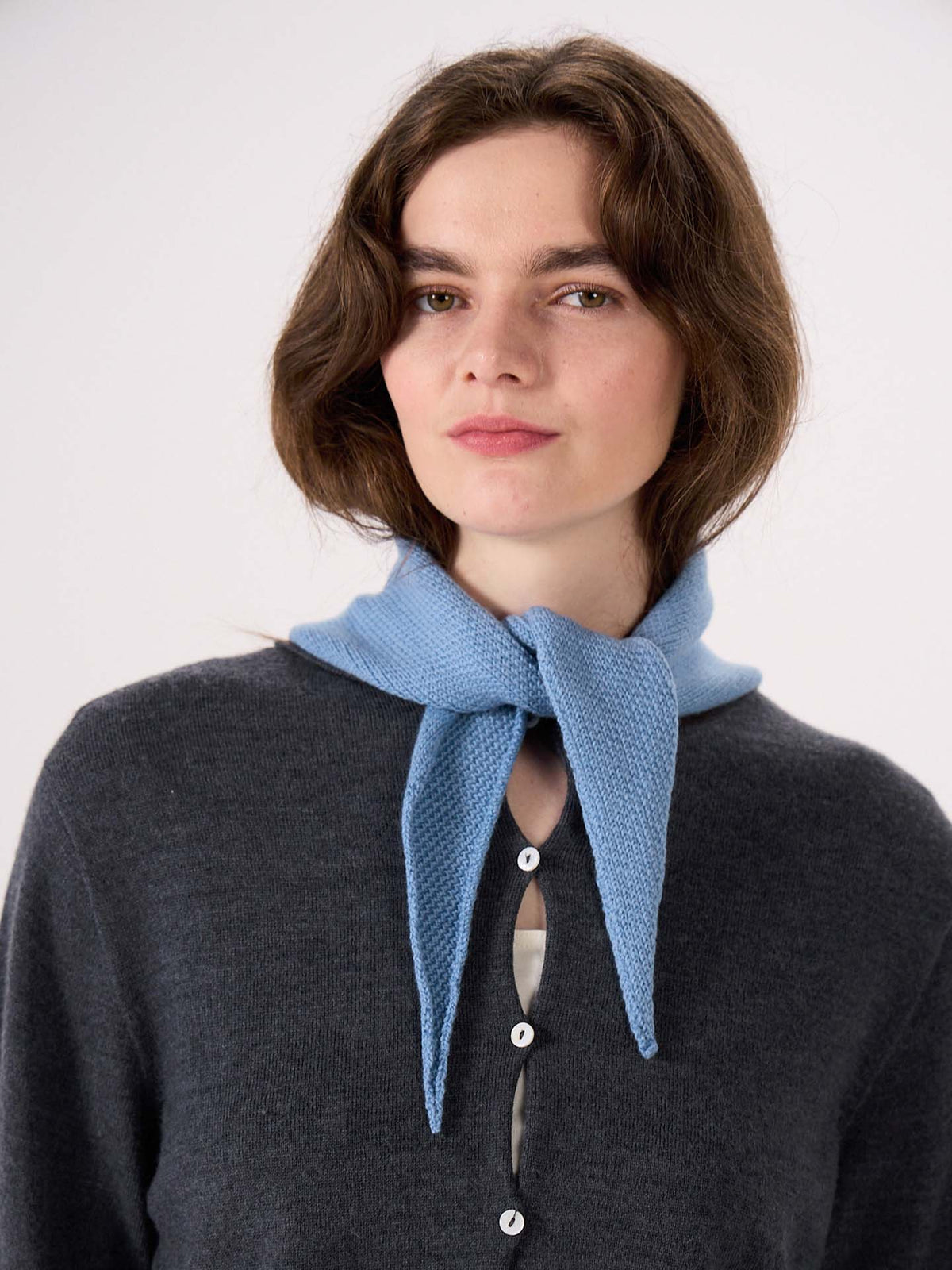 A person wearing a dark grey button-up sweater and the Daisy Scarf in Celeste Blue—measuring 97cm long and 22cm wide—tied around their neck, standing against a plain background.