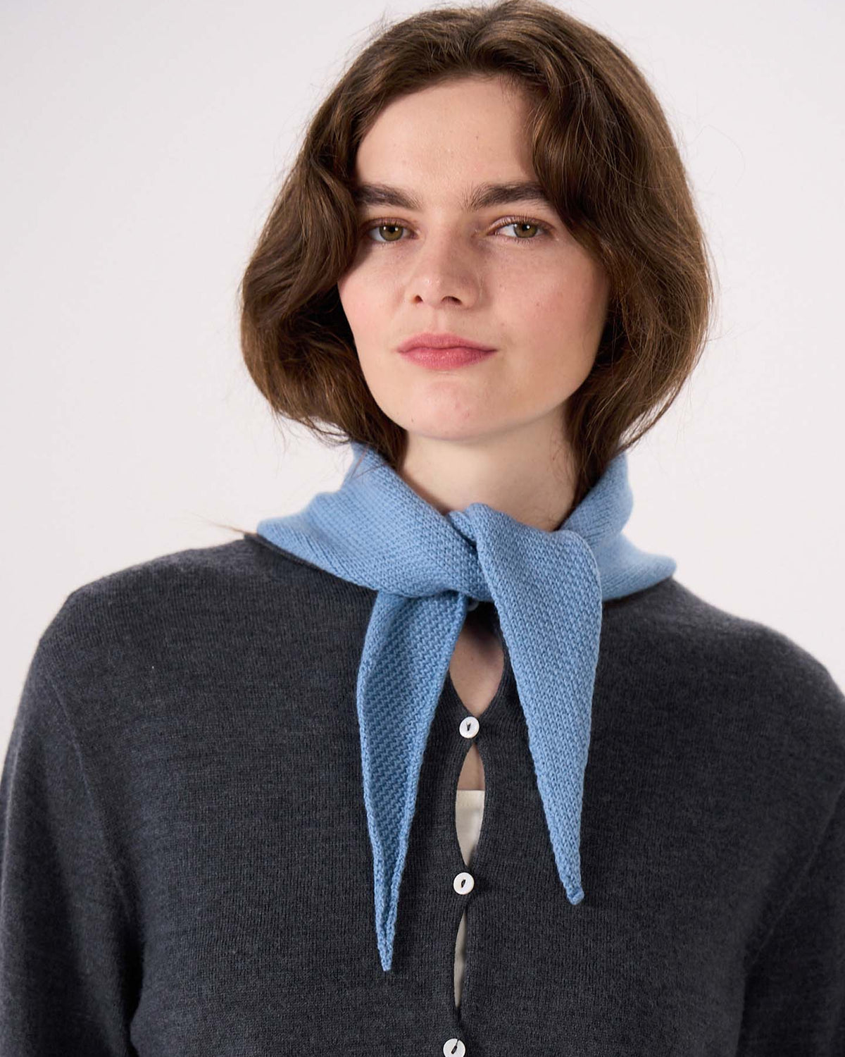 A person wearing a dark grey button-up sweater and the Daisy Scarf in Celeste Blue—measuring 97cm long and 22cm wide—tied around their neck, standing against a plain background.