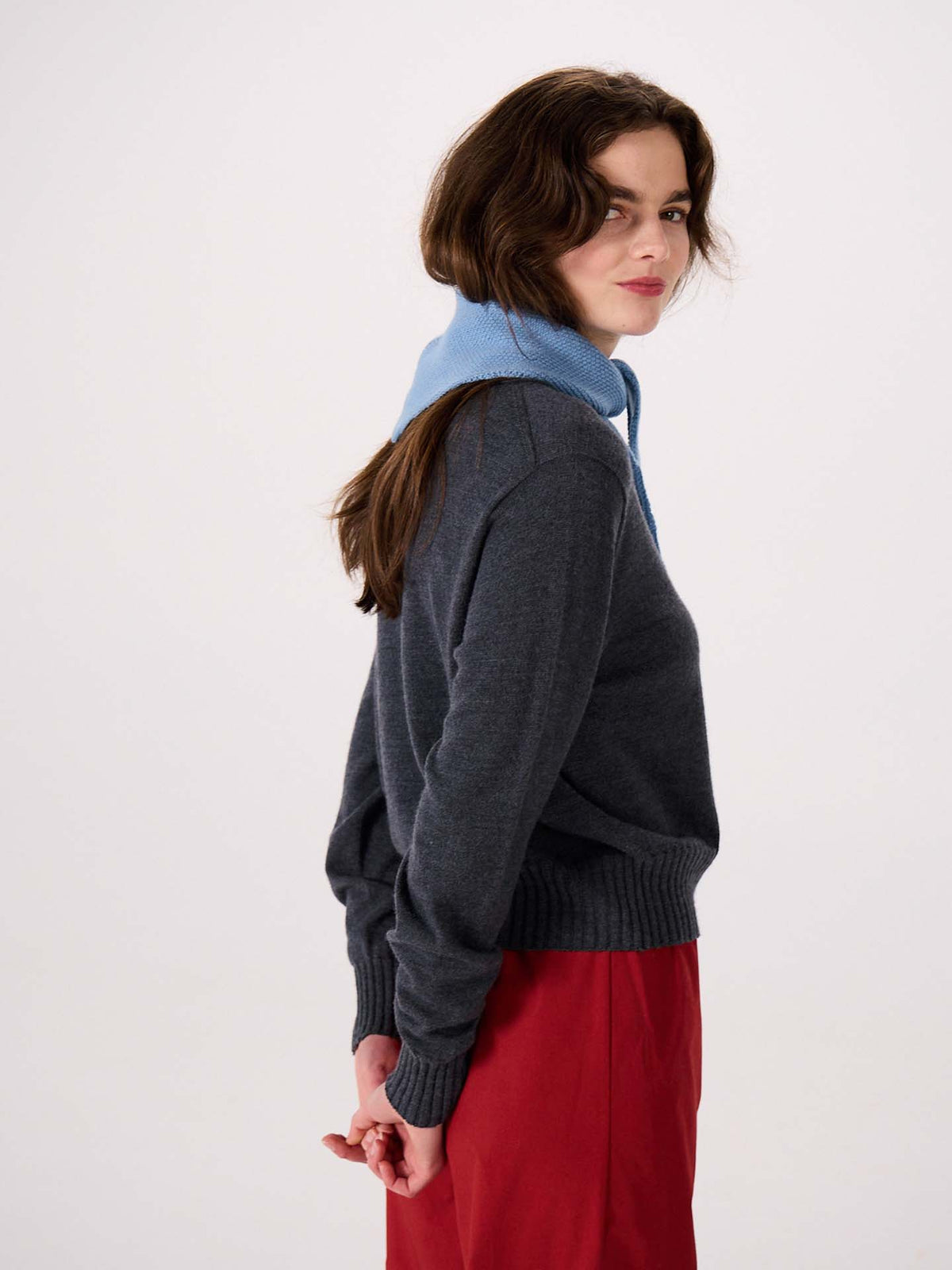 A woman with wavy brown hair stands sideways against a plain background, wearing a dark gray sweater and red skirt. She has the Daisy Scarf in Celeste Blue, measuring 22cm wide and 97cm long, draped over her shoulders with her hands behind her back.