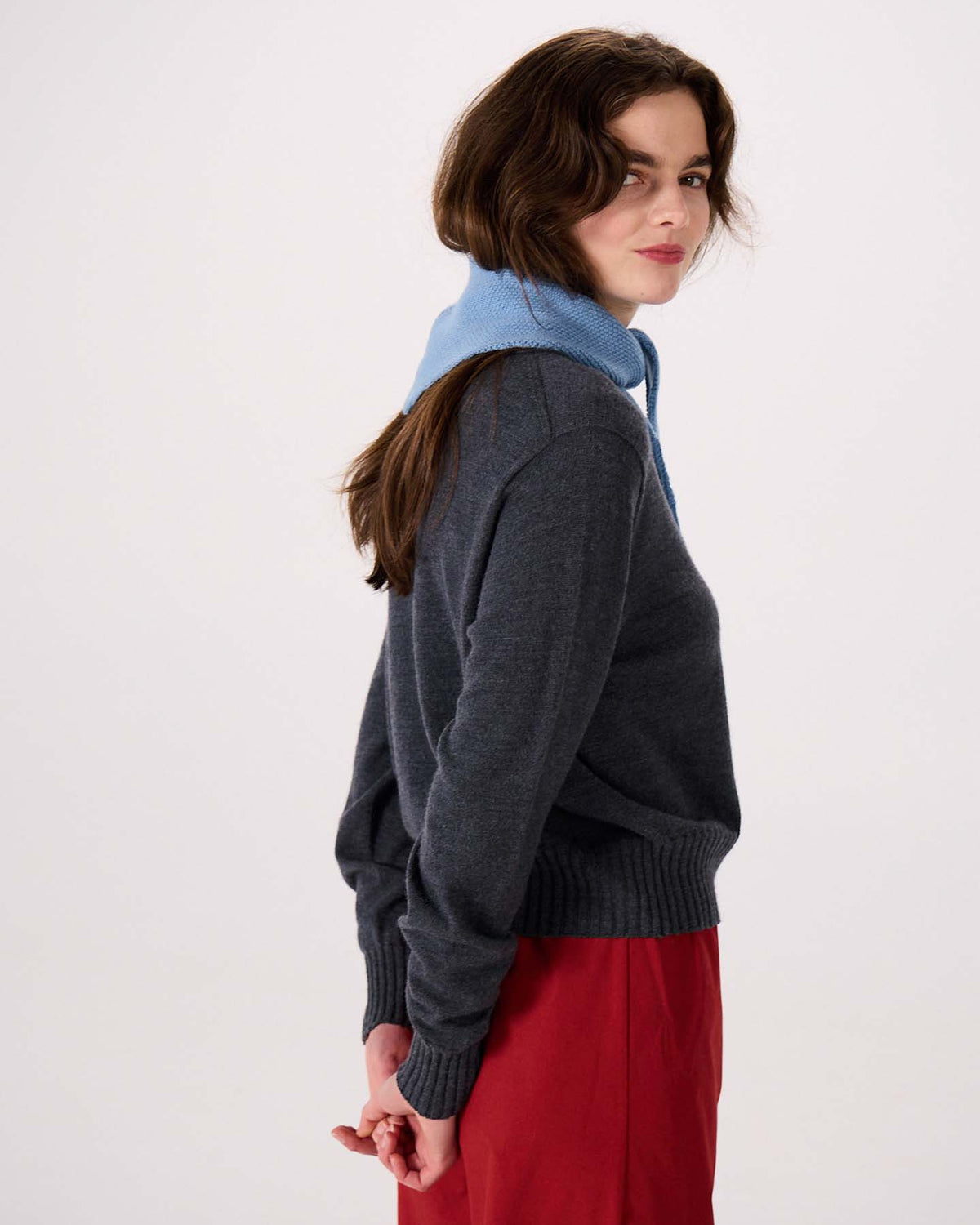 A woman with wavy brown hair stands sideways against a plain background, wearing a dark gray sweater and red skirt. She has the Daisy Scarf in Celeste Blue, measuring 22cm wide and 97cm long, draped over her shoulders with her hands behind her back.